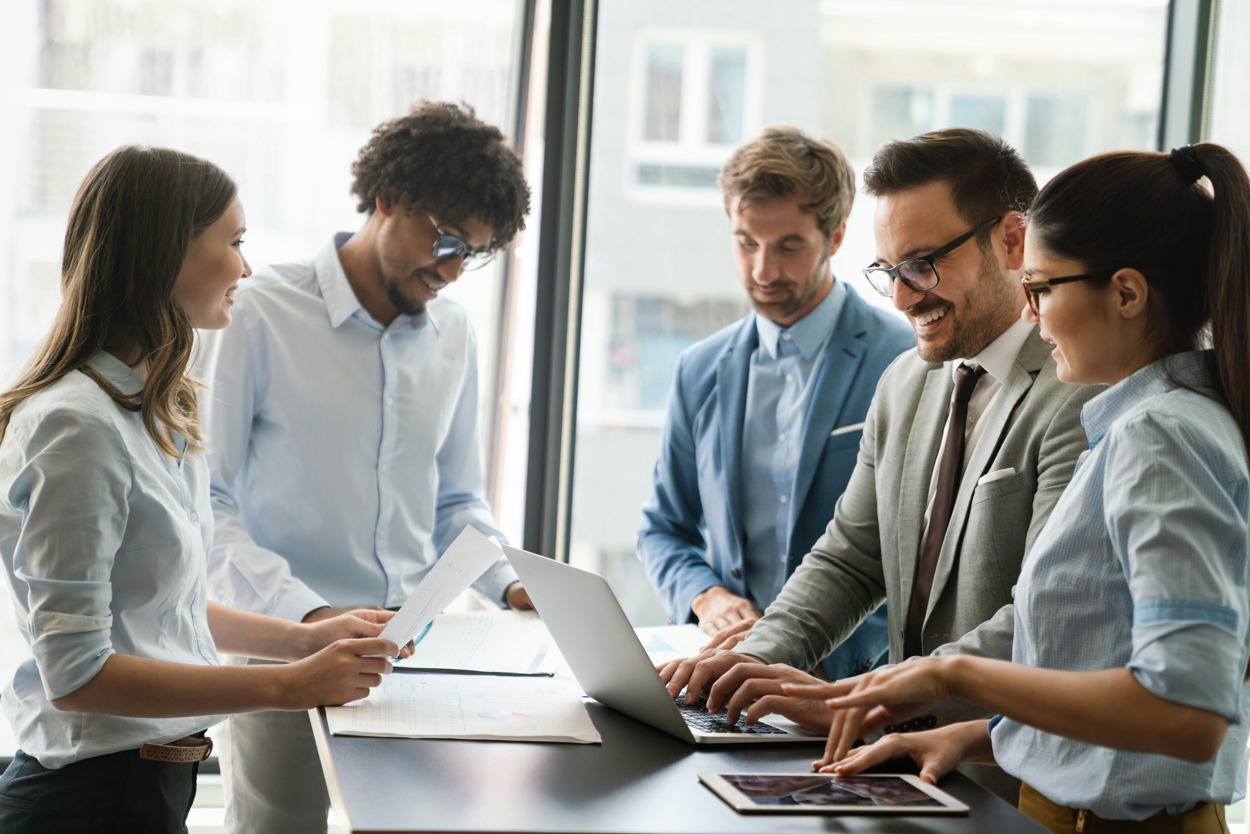 Group of five diverse professionals working together in a modern office, using laptops and documents, smiling and collaborating.