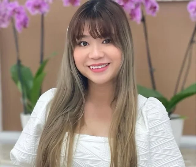A smiling young woman with long, wavy blonde hair and bangs, wearing a white top, standing in front of a background with pink orchids and green leaves.