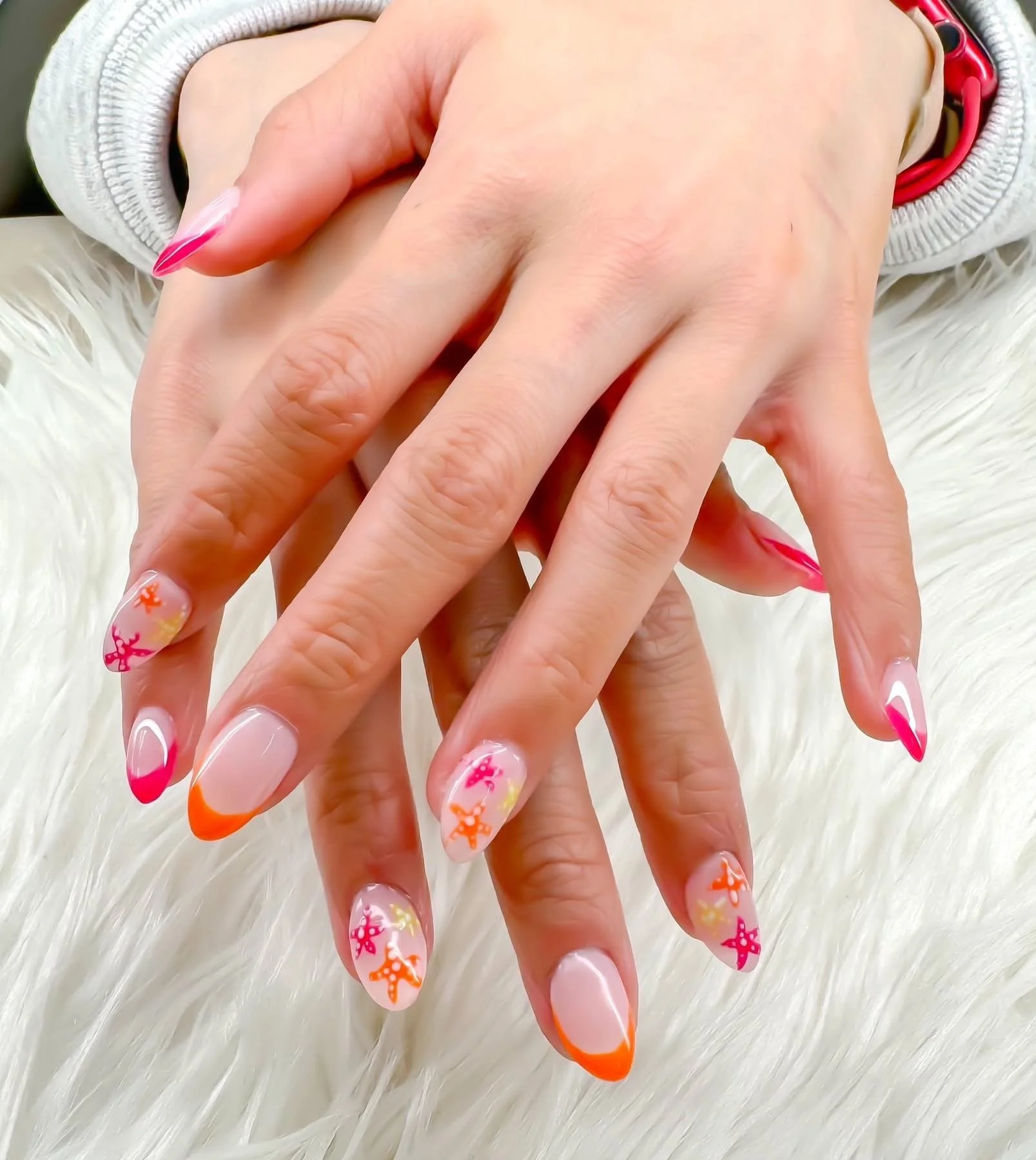 Close-up of a customer's hands with decorated nails featuring a pink and orange design with floral and butterfly patterns, resting on a fluffy white surface.