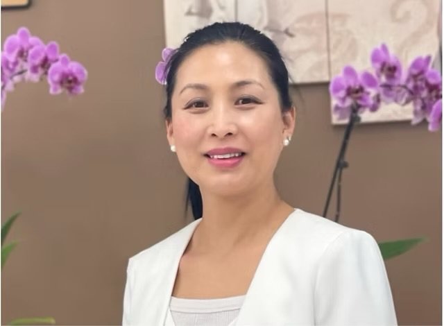 A woman with black hair tied back, wearing a white top and pearl earrings, smiling in front of purple orchids and a neutral background.