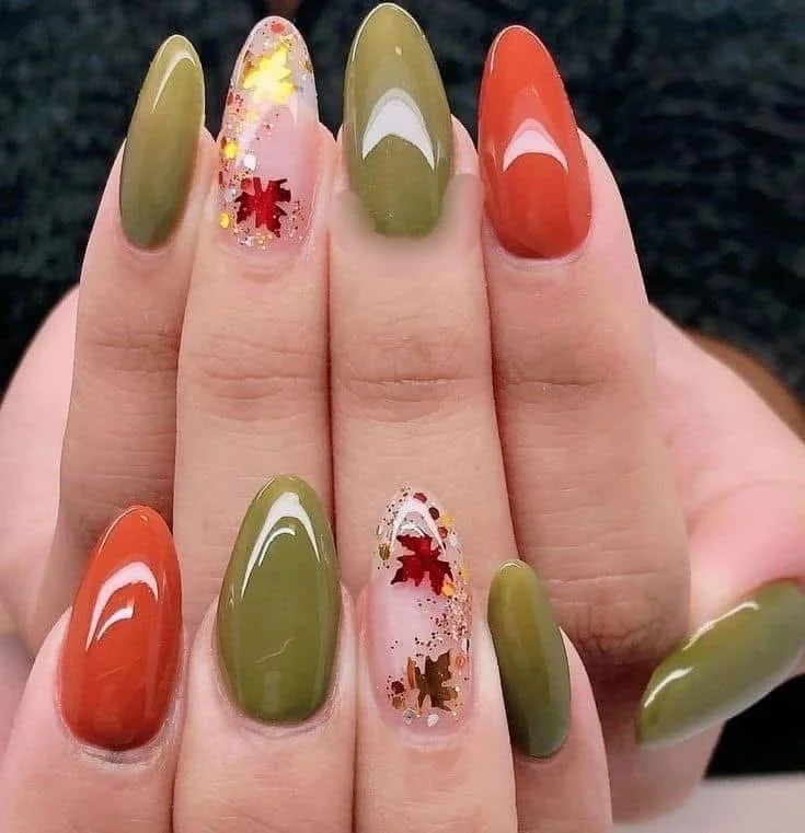 Close-up of a hand showing nails painted with autumn-themed nail art, with some nails in olive green, others in orange, and two accent nails decorated with clear polish and red and gold maple leaf confetti.