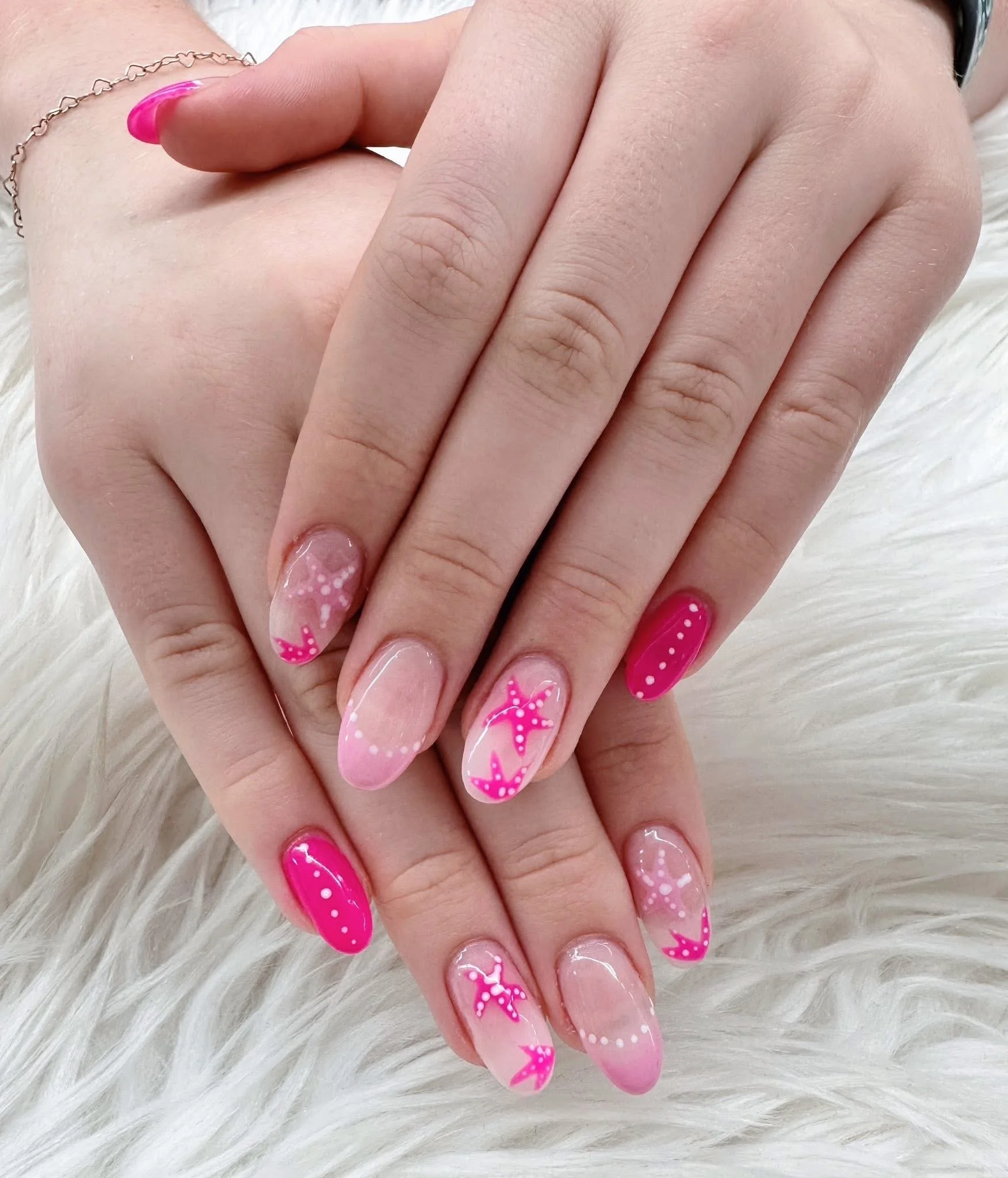 Close-up of hands with pink and clear stiletto nails decorated with white and pink starfish and dots, resting on a white fur surface.