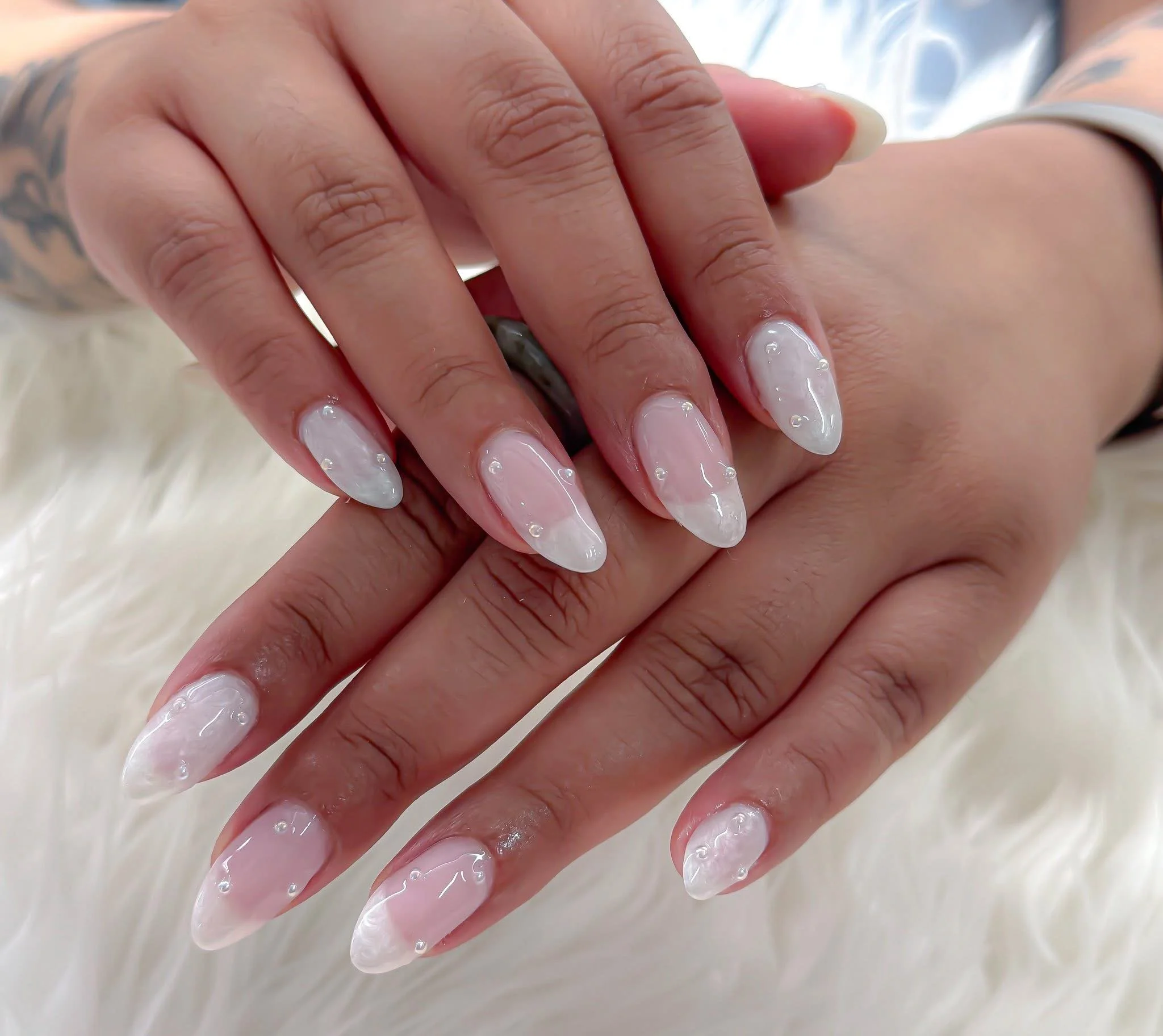 Close-up of hands with manicured nails featuring a white and pearl design with small pearl accents, resting on a white furry surface.
