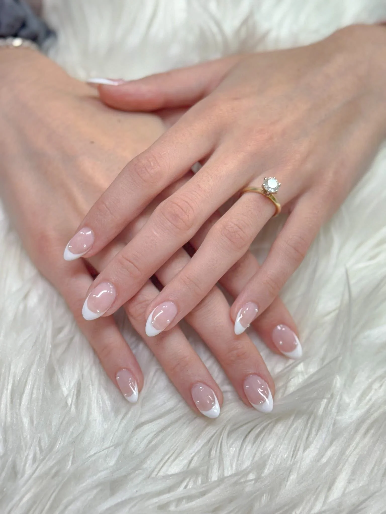 Close-up of a customer's hands resting on a soft, white, furry surface. The woman has manicured nails with a French tip design and is wearing a gold ring with a single round gemstone on her ring finger.