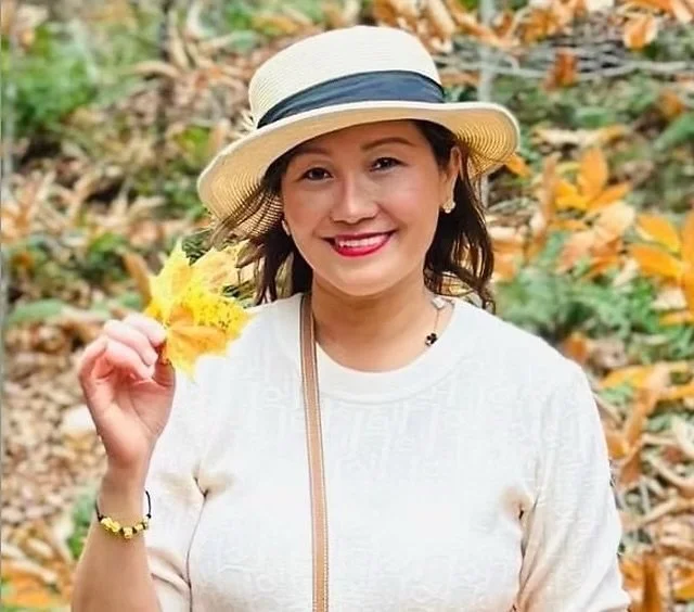 A woman wearing a cream-colored sweater and a straw hat, holding yellow leaves in her right hand, smiling outdoors with fallen autumn leaves on the ground.