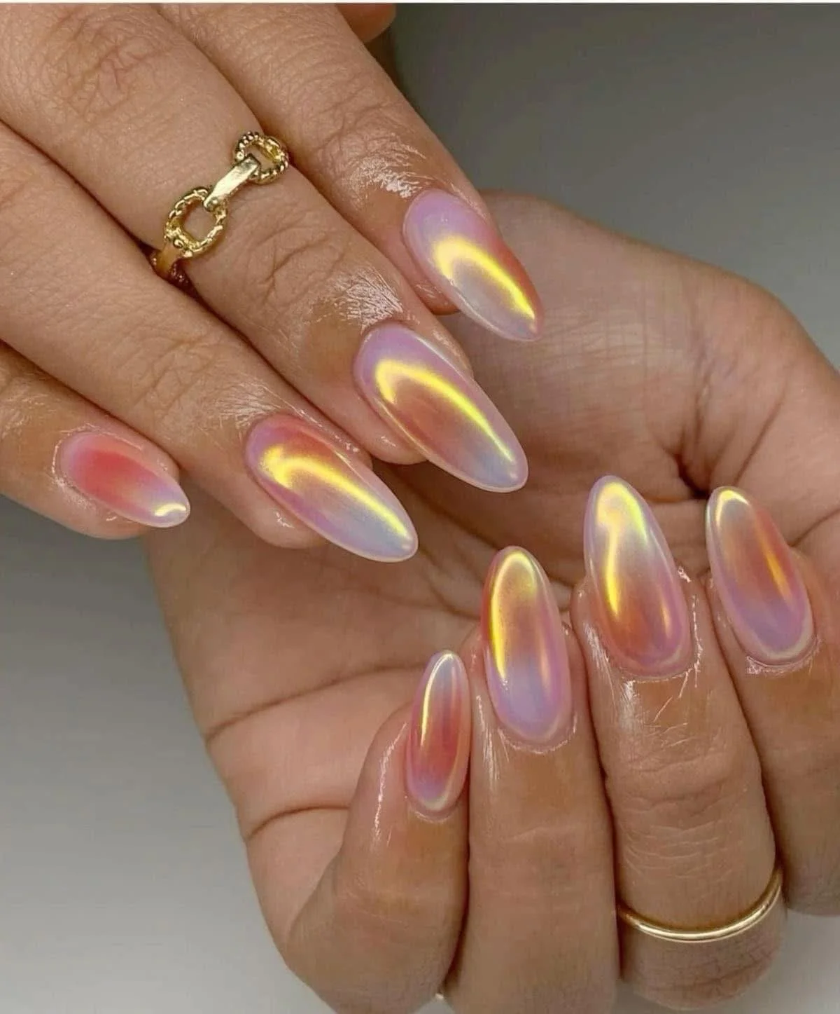 Close-up of a hand with long, oval-shaped, iridescent nail art featuring a mirror-like finish in pastel hues, and a gold ring on the finger.