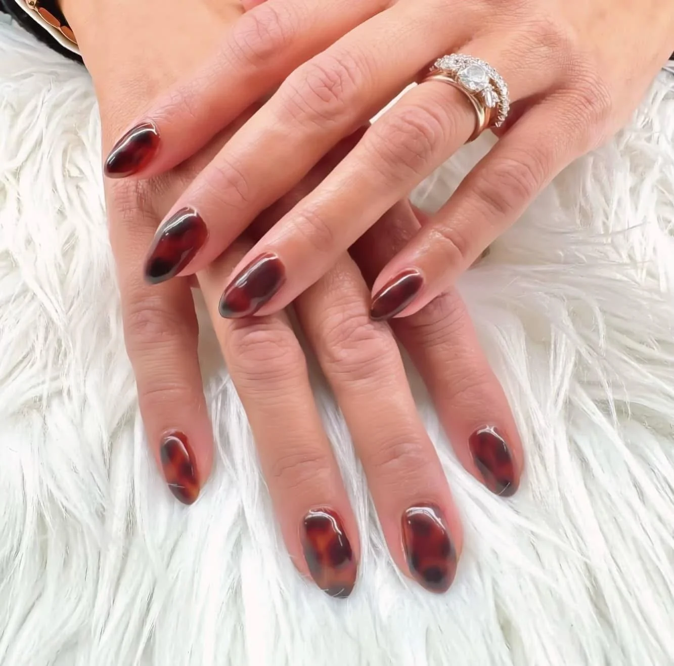 Close-up of hands with tortoise shell nail polish, adorned with a large engagement ring with a central diamond, resting on a fluffy white surface.