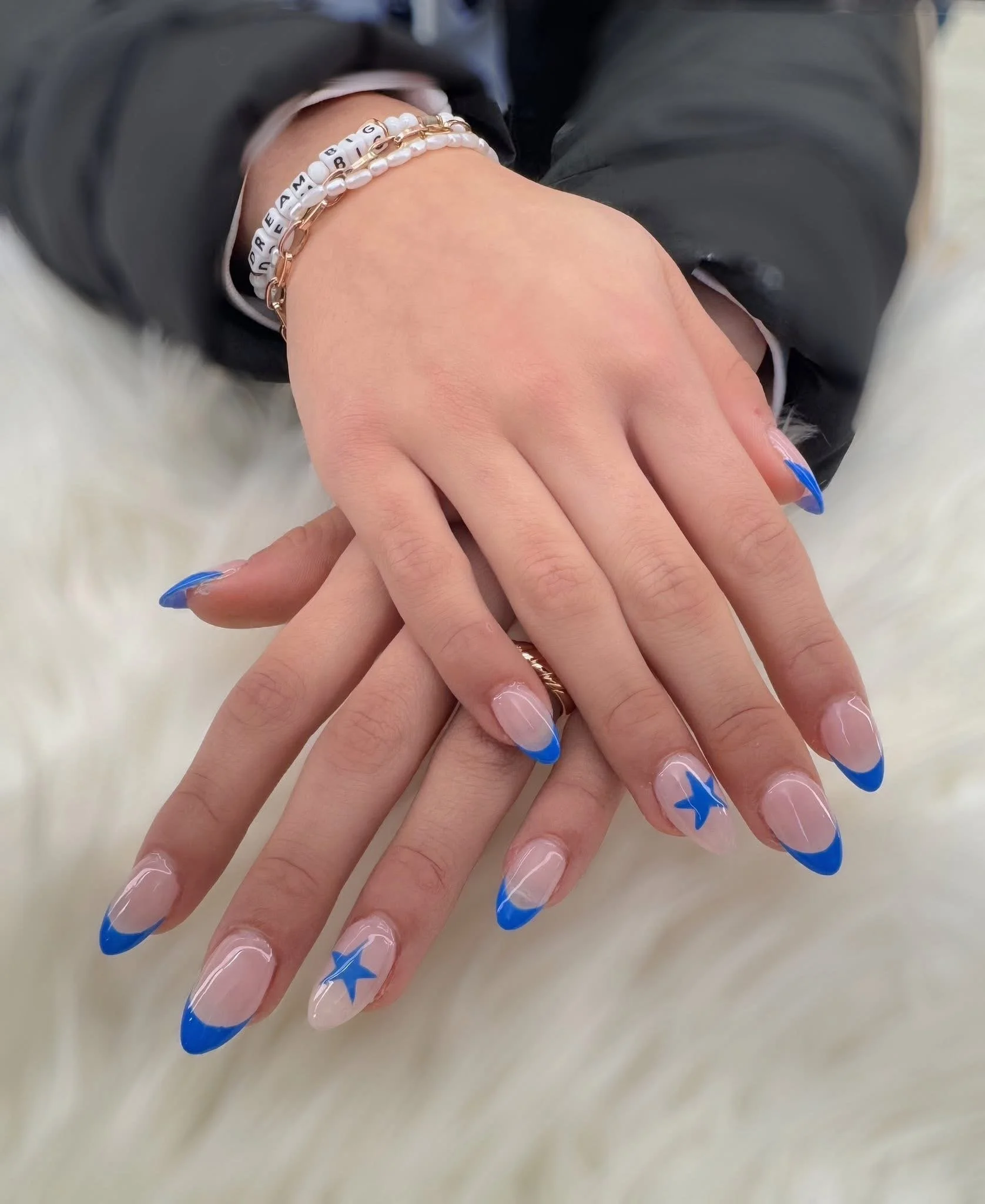 Close-up of manicured nails with blue French tips and star designs, jewelry on fingers, and a person's hand resting on a white furry surface.