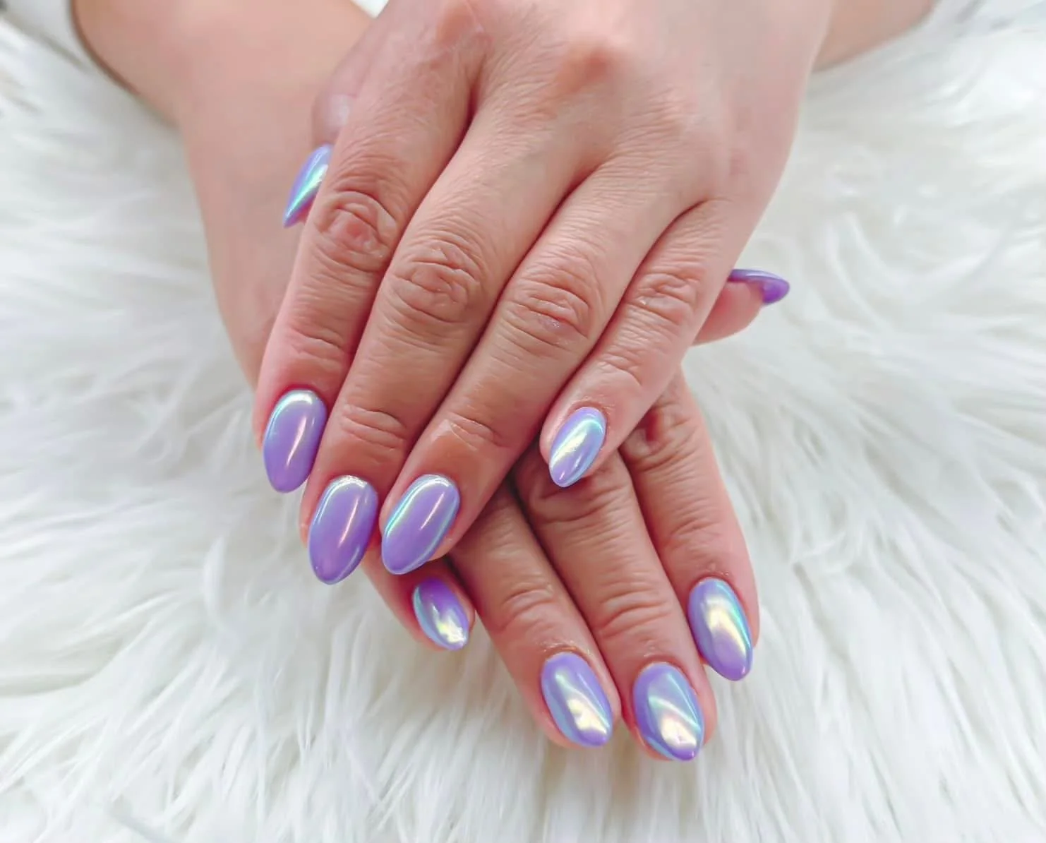 Close-up of hands with iridescent purple and blue oval-shaped nails resting on a fluffy white surface.