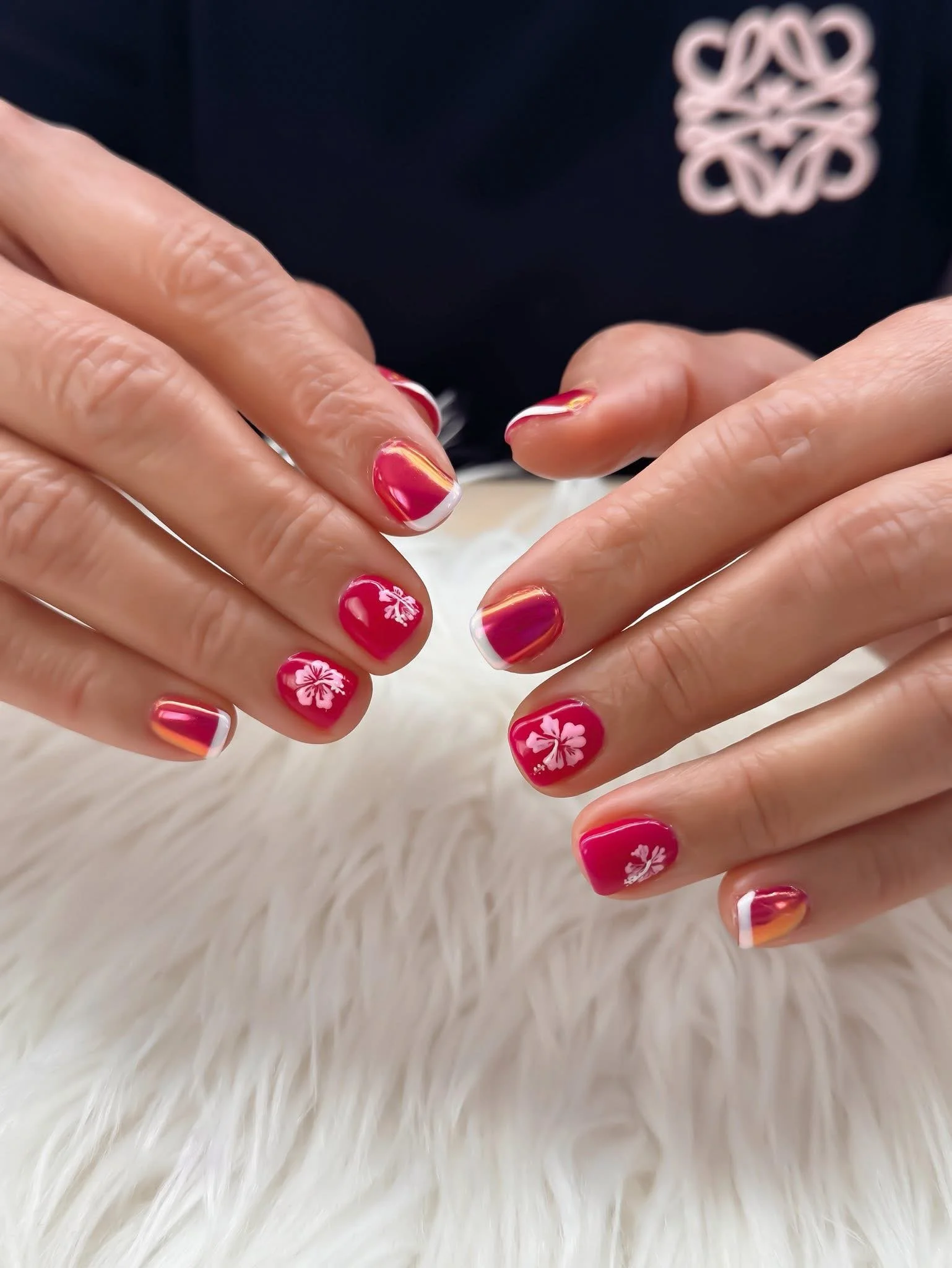 Close-up of hands with red and gold decorated nails, featuring white floral designs and French tips, resting on a white furry surface.