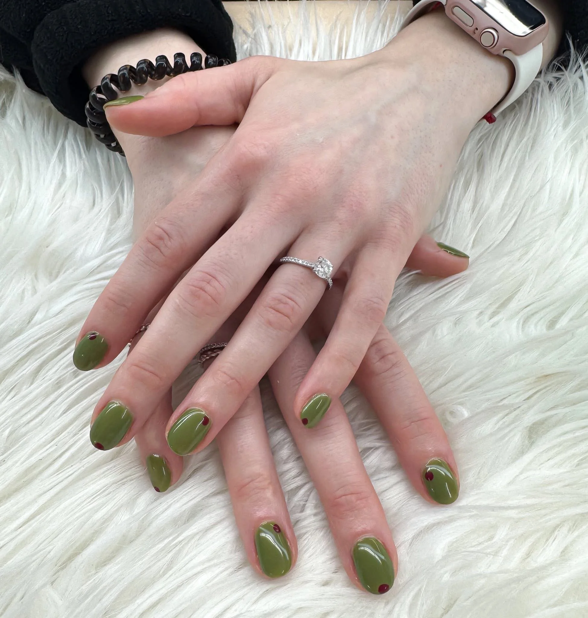 Close-up of two hands with olive green nail polish and small red dots on each nail, resting on a white furry surface. One hand has a diamond engagement ring and a black beaded bracelet, the other hand has a smartwatch.