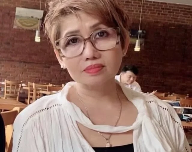 A woman with short, light brown hair and glasses, wearing a white top, sitting in a restaurant with wooden furniture and brick walls, with other patrons in the background.
