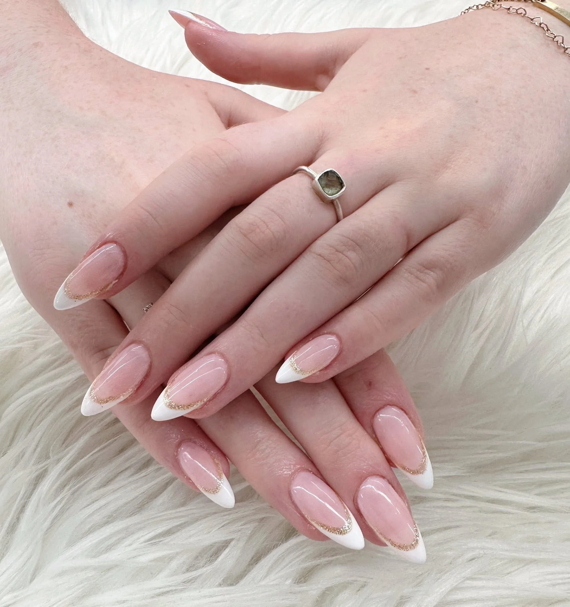 Close-up of hands with manicured nails resting on a fluffy white surface. The nails have a French tip design with white tips, nude pink bases, and gold glitter accents. One hand wears a silver ring with a green gemstone, and a delicate chain bracelet is visible.