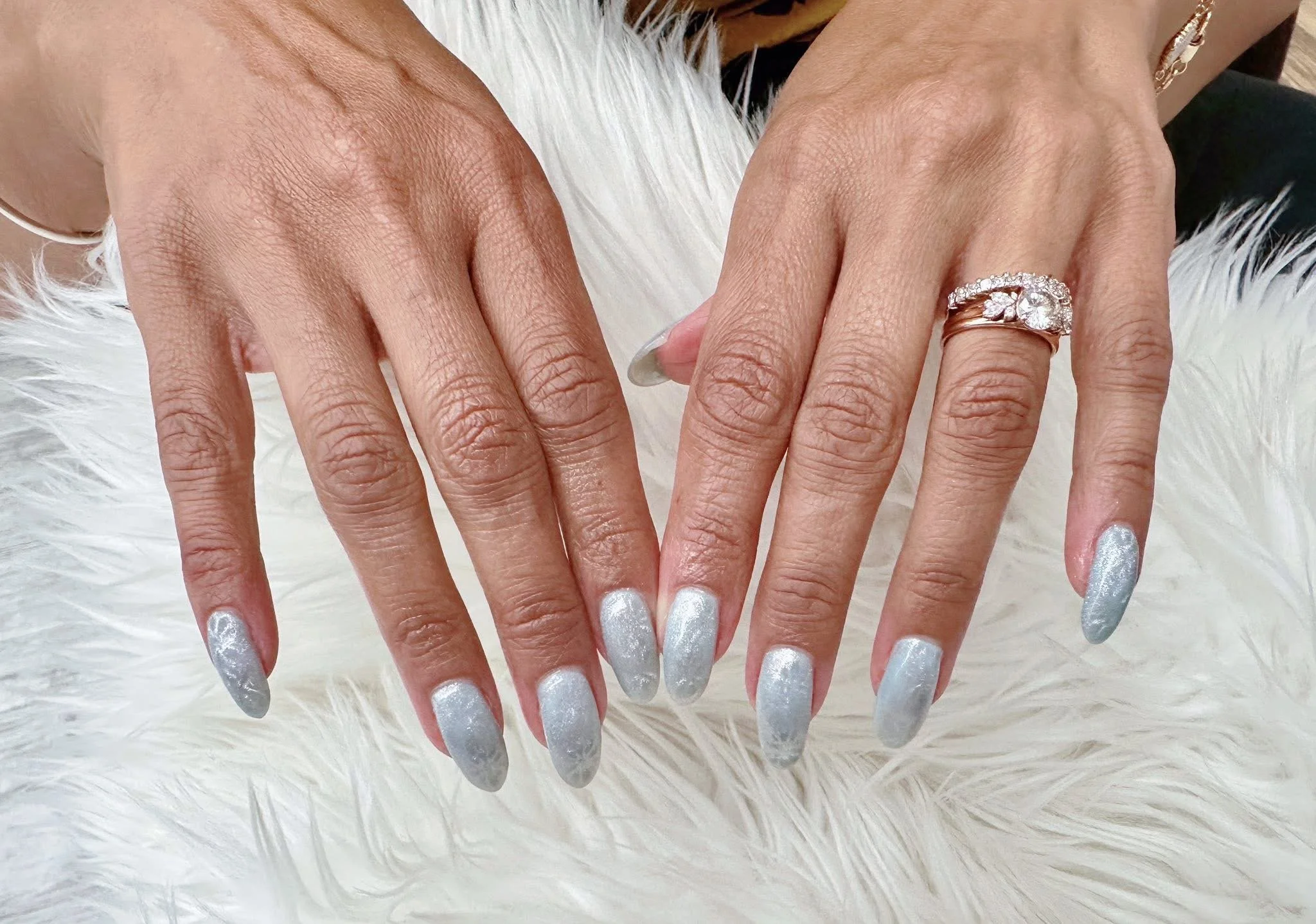 A pair of hands with manicured nails painted in a shimmering light blue color, resting on a white furry surface. One hand is adorned with a large diamond engagement ring and a wedding band.