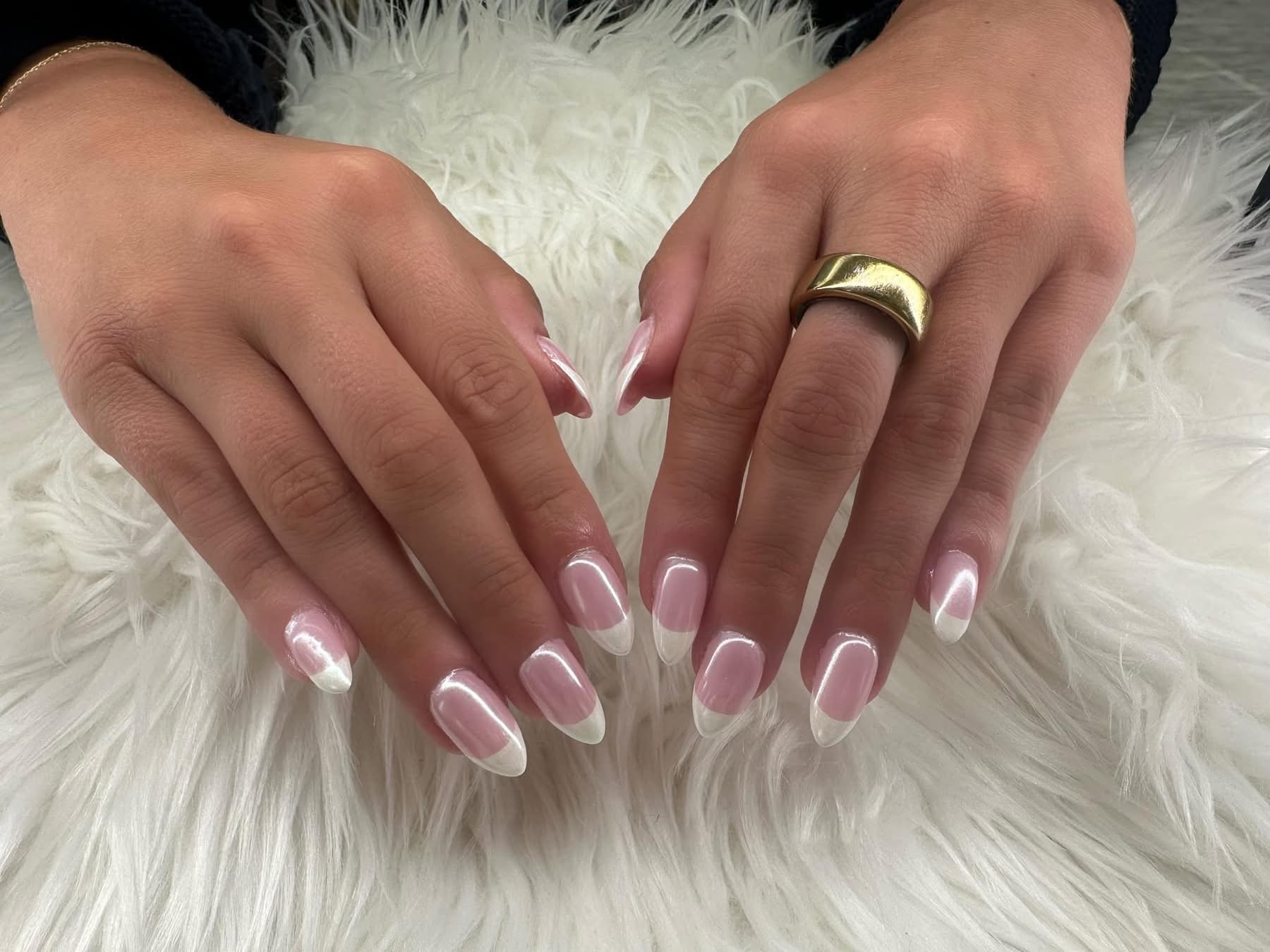 A pair of hands with French manicured nails resting on a white furry surface, one hand has a gold ring on the ring finger.