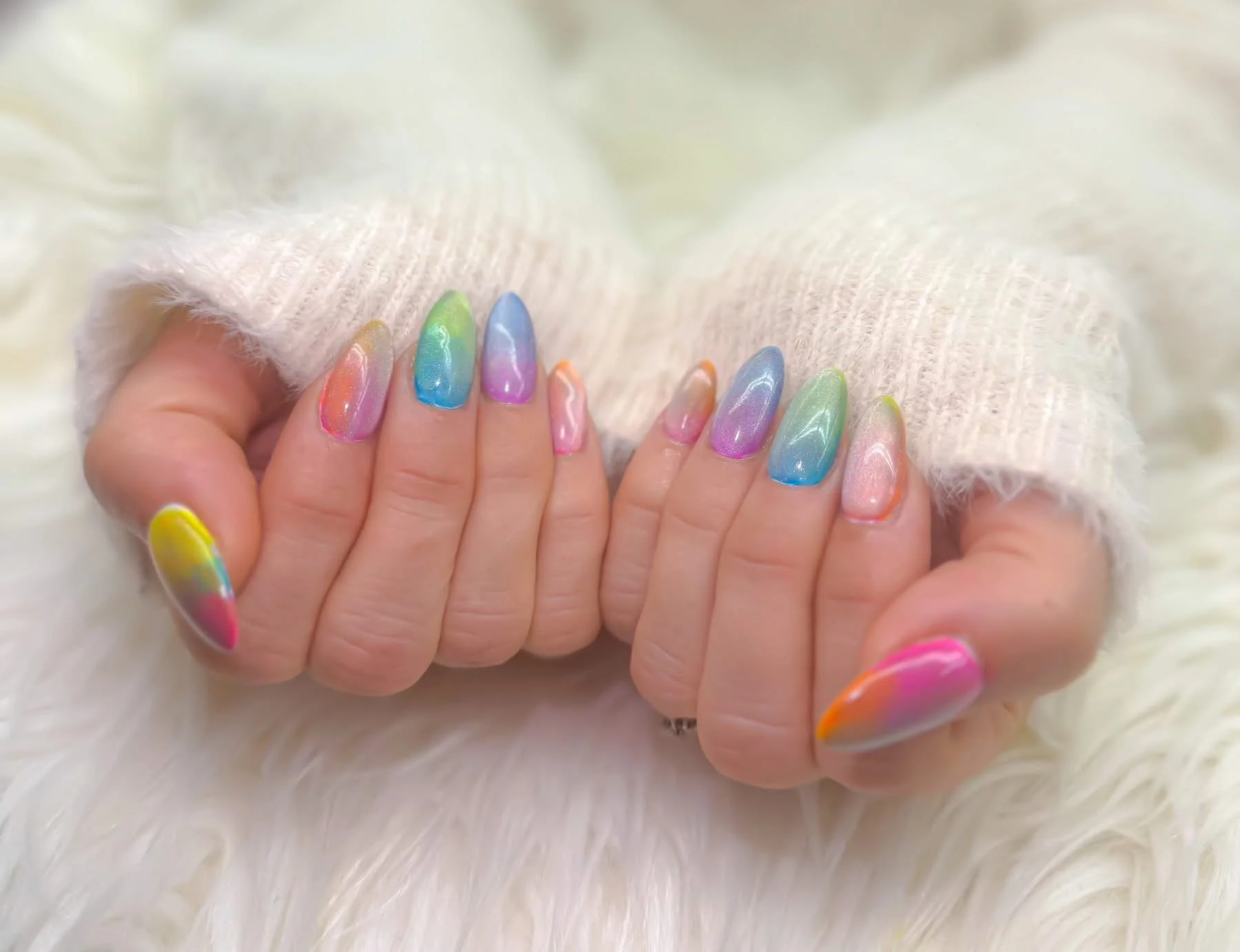 Hand with rainbow-colored, tie-dye style nail polish, resting on a soft, fuzzy white surface, with a cream-colored knit sweater partially visible.