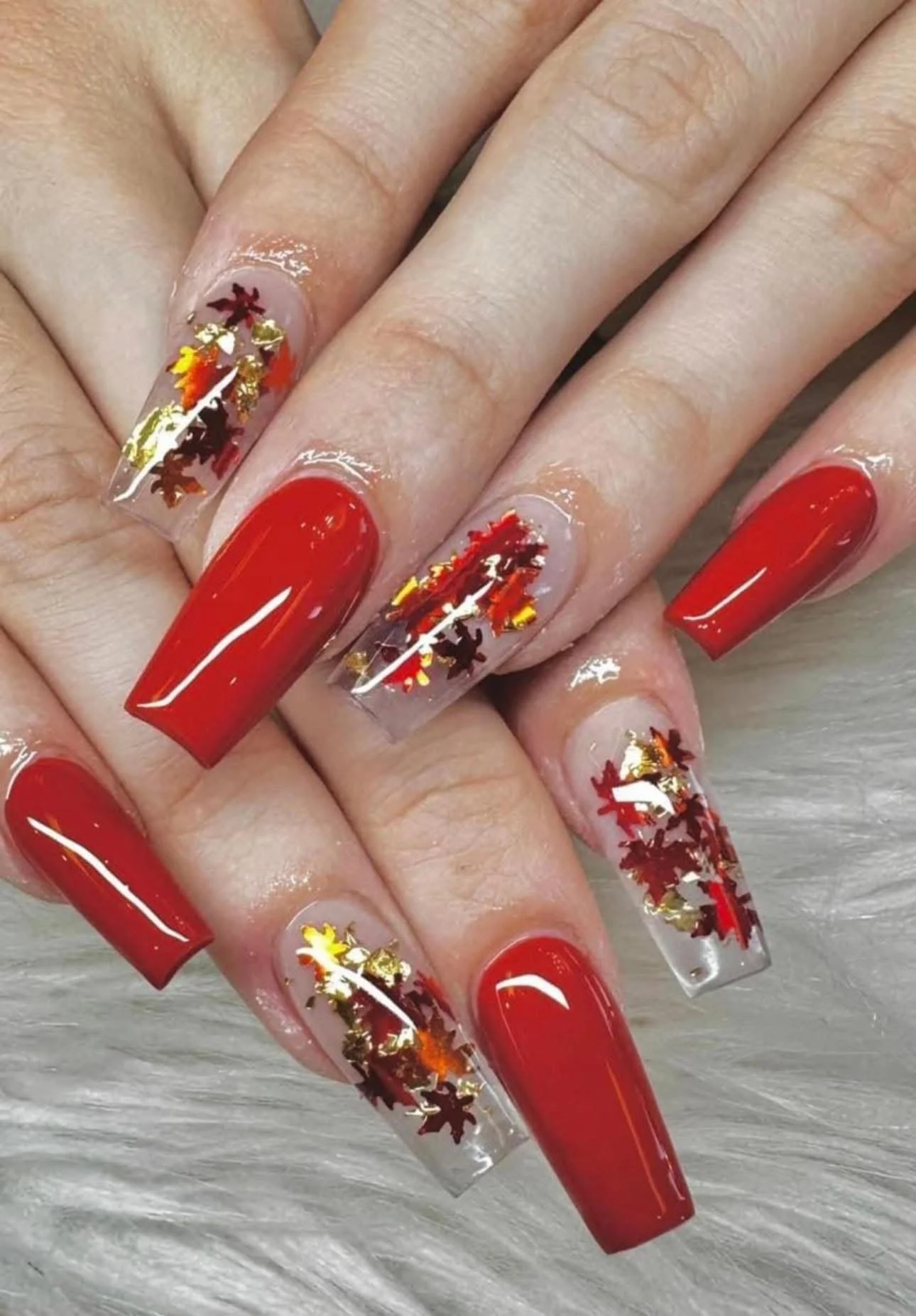 Nail art with red and clear nails, decorated with red, orange, yellow, and gold leaf-shaped glitter inside the clear nails.
