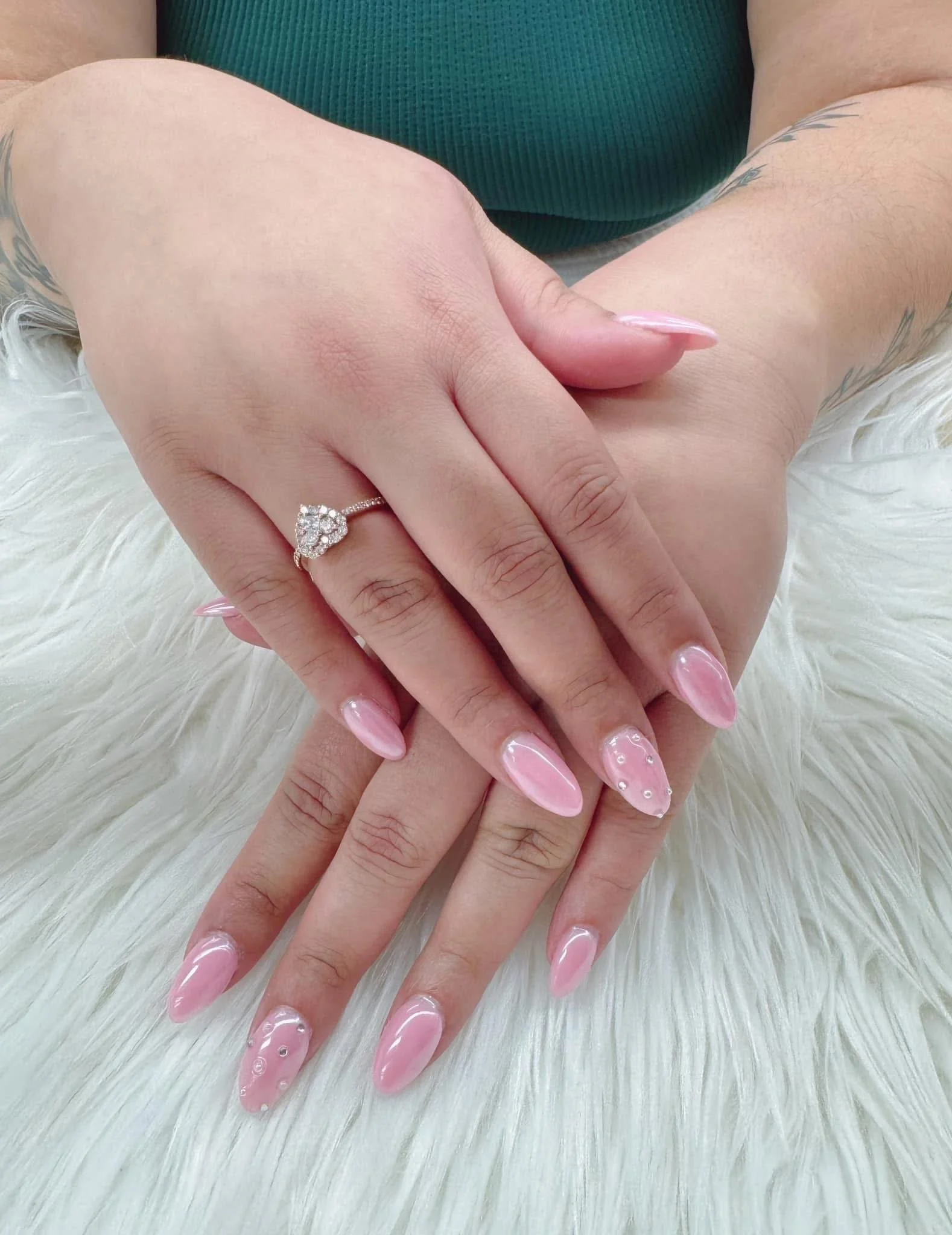 Hands with pink manicured nails, one hand displaying a diamond engagement ring with a heart-shaped design, resting on a white furry surface.