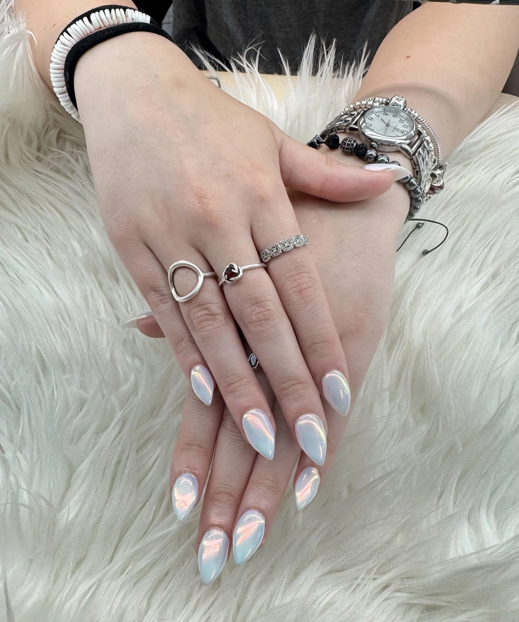Close-up of hands resting on a white, fur-like surface, adorned with silver rings, a watch, and multiple bracelets, with iridescent almond-shaped nails.