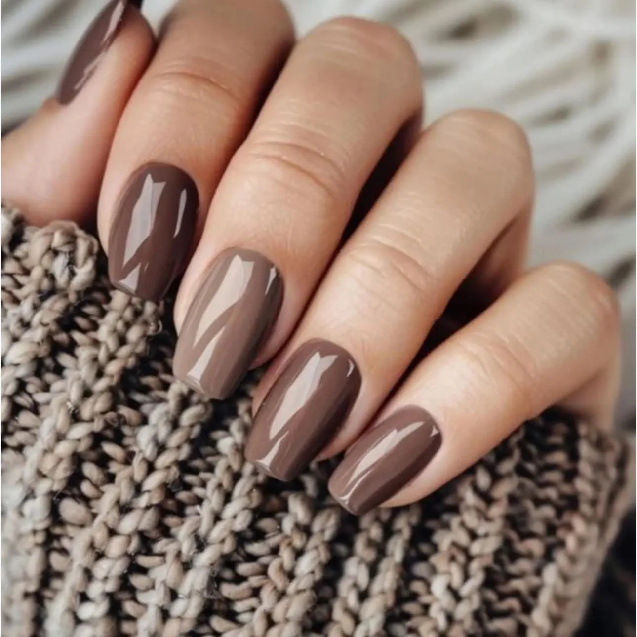 Close-up of a person's hand with neatly manicured nails painted in a taupe or beige color, resting on a textured knitted fabric.