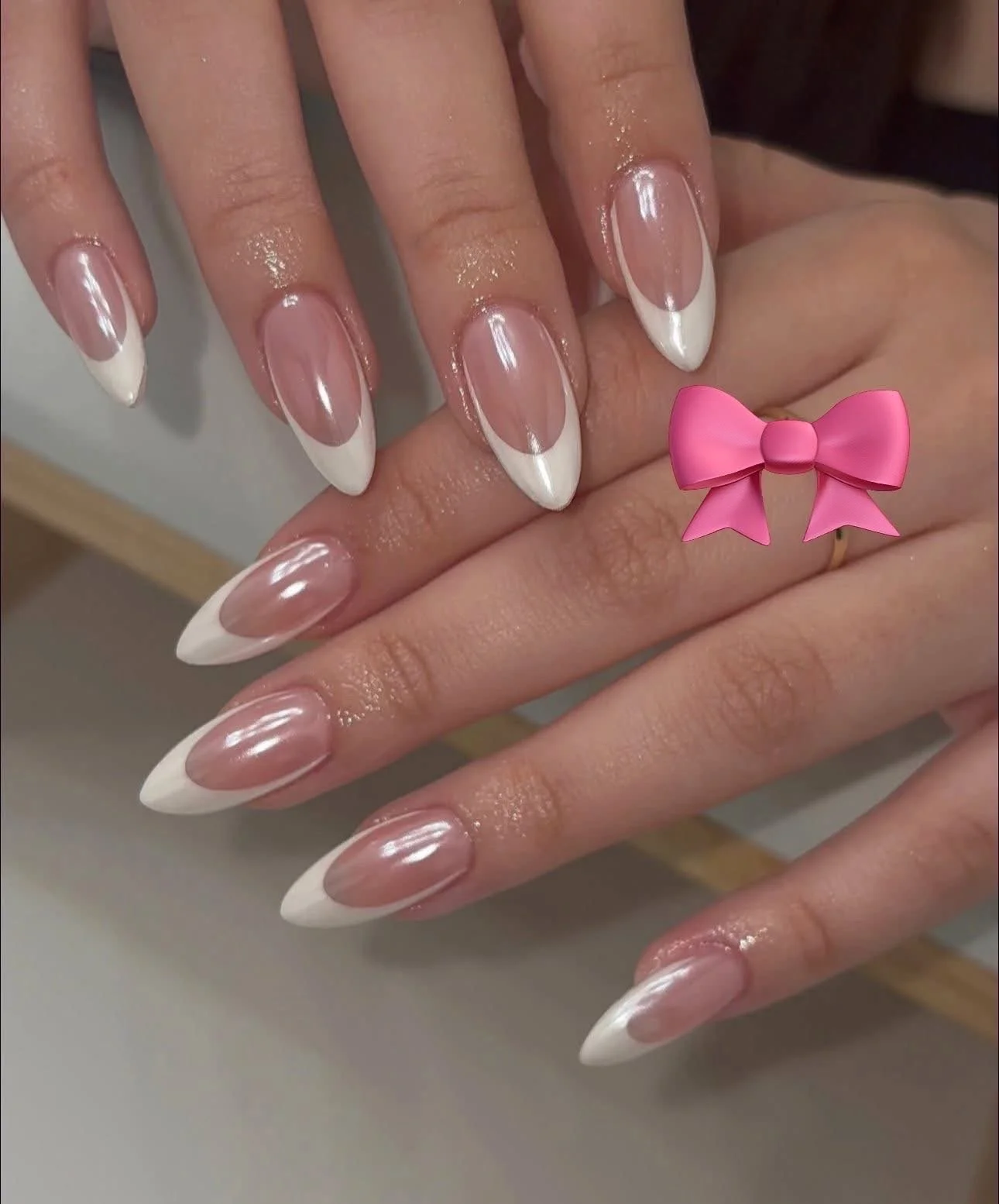 Close-up of hands with manicured nails featuring a clear base and white French tips, with a pink bow sticker added to the image.