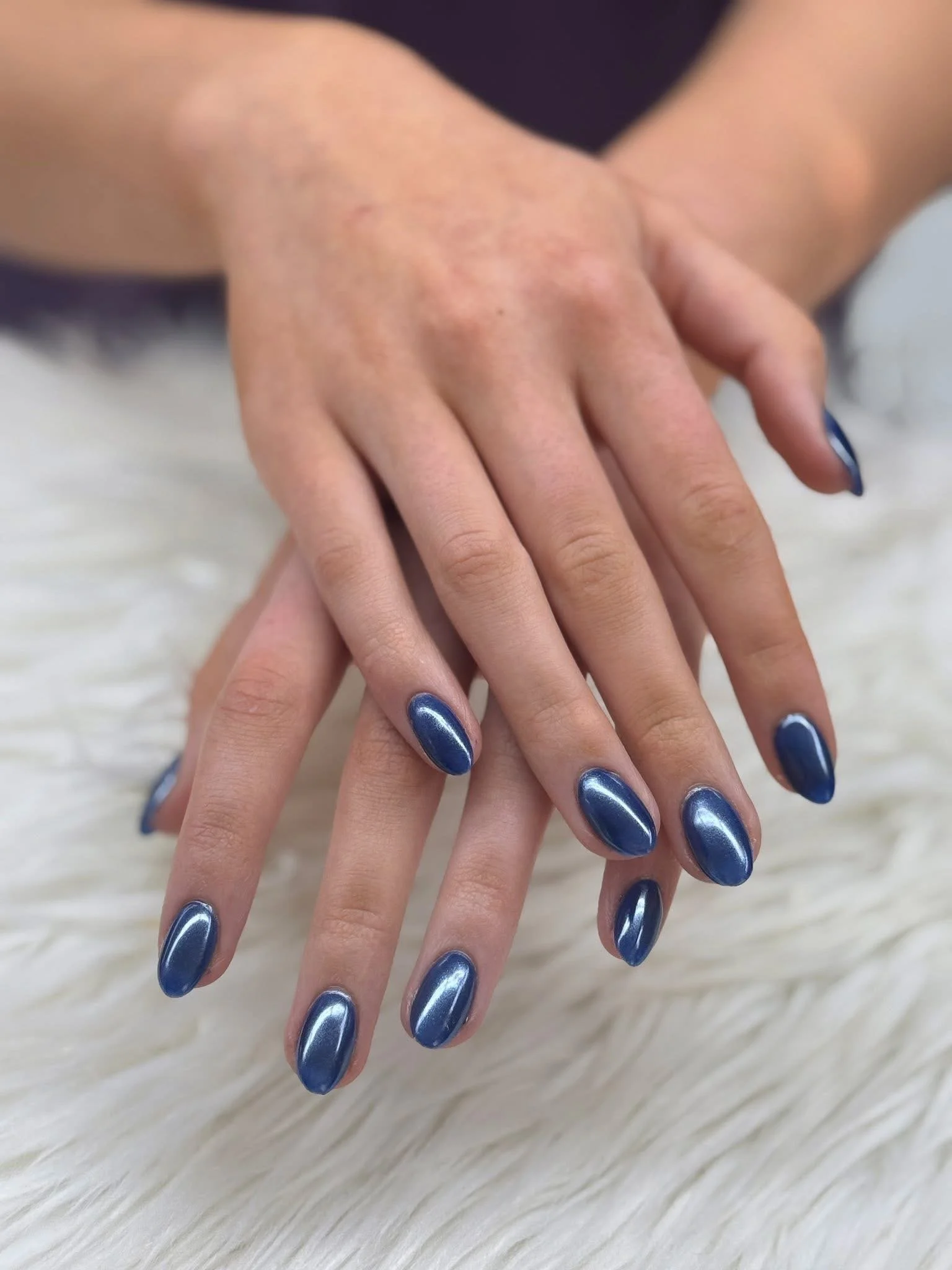 Hands with shiny dark blue painted nails resting on a soft white surface.