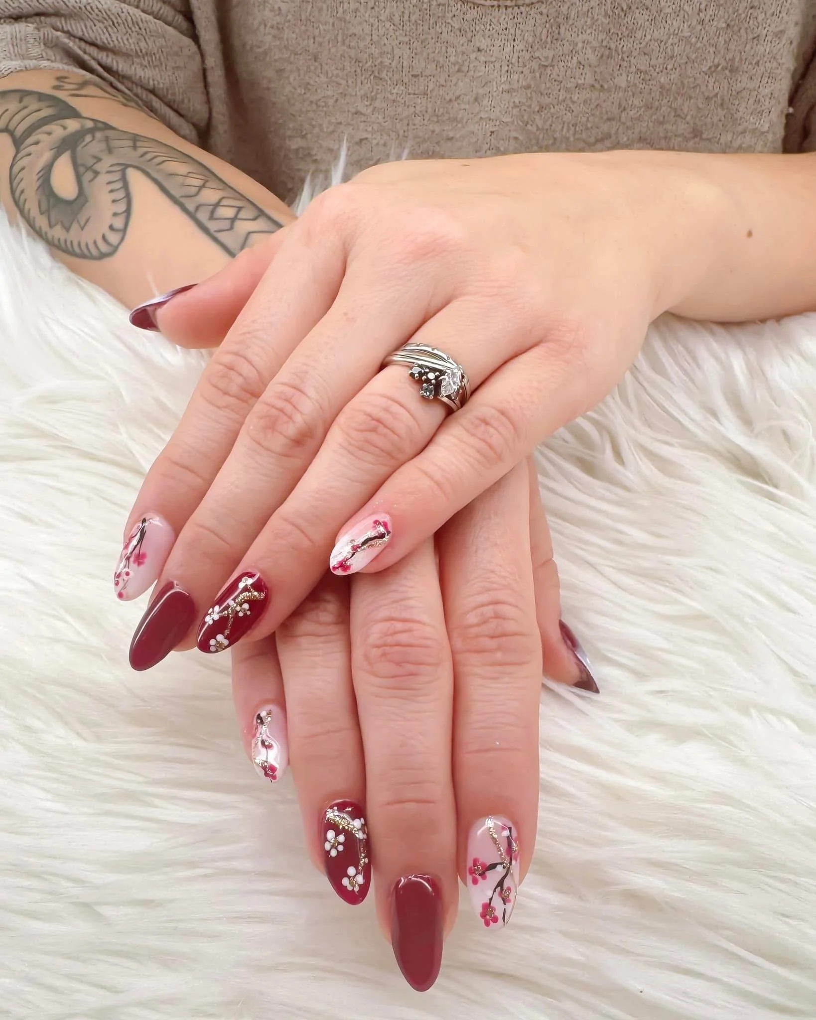 Close-up of hands with decorated nails resting on a white furry surface, showing a large diamond ring on the ring finger, and a tattoo on the arm.