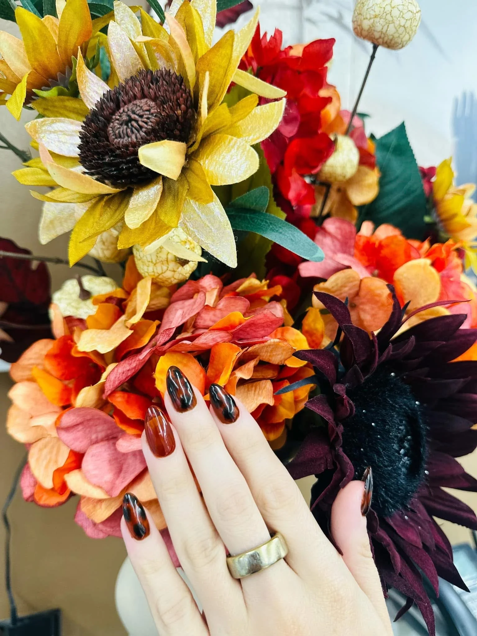 Close-up of an artificial floral arrangement with sunflowers, daisies, and other colorful flowers, held by a hand with painted nails and a gold ring.