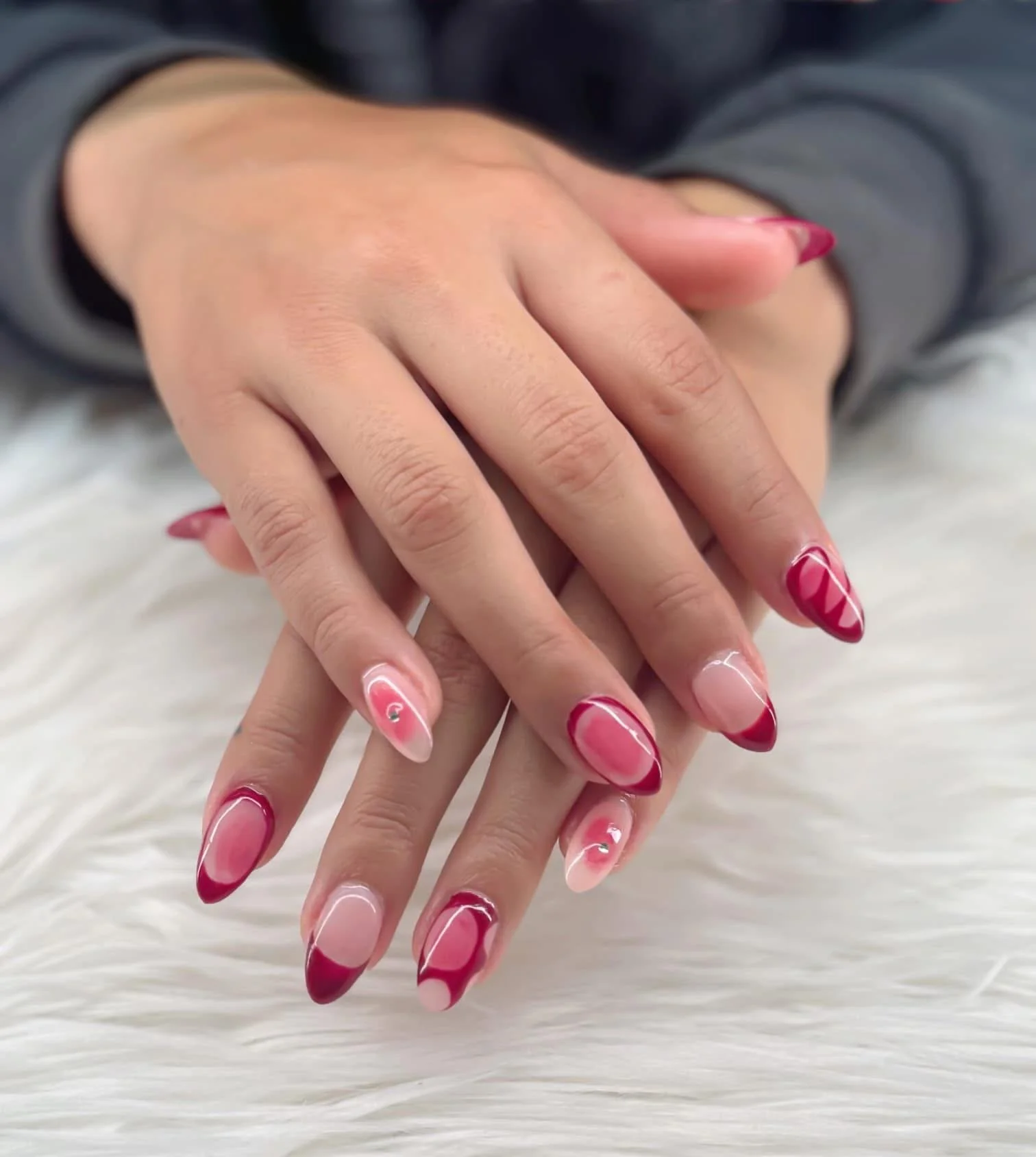 Close-up of hands with manicured nails featuring pink and red swirl nail art, resting on a white furry surface.