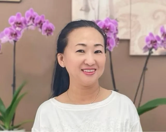 Smiling woman with dark hair wearing a white shirt, standing in front of pink orchids and wall art.