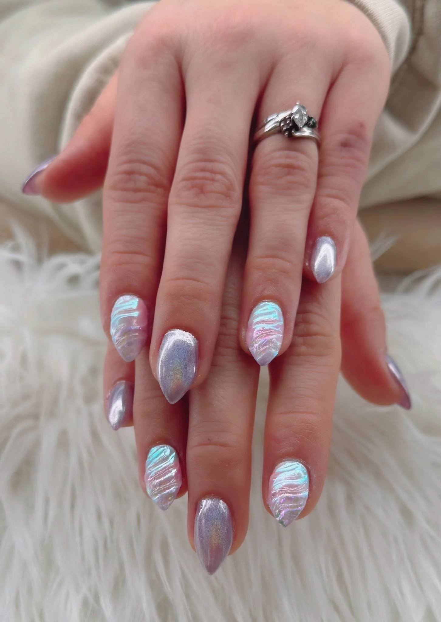 Close-up of a customer's hands with holographic and silver metallic nail art, and a silver ring on her middle finger, resting on a white, furry surface.