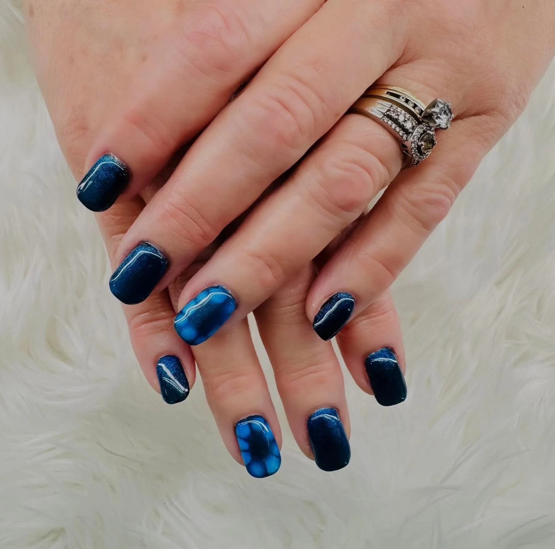 Close-up of a customer's hand with a blue and black marble design on their nails, wearing multiple rings including a wedding band and a ring with a skull motif, against a light-colored furry background.