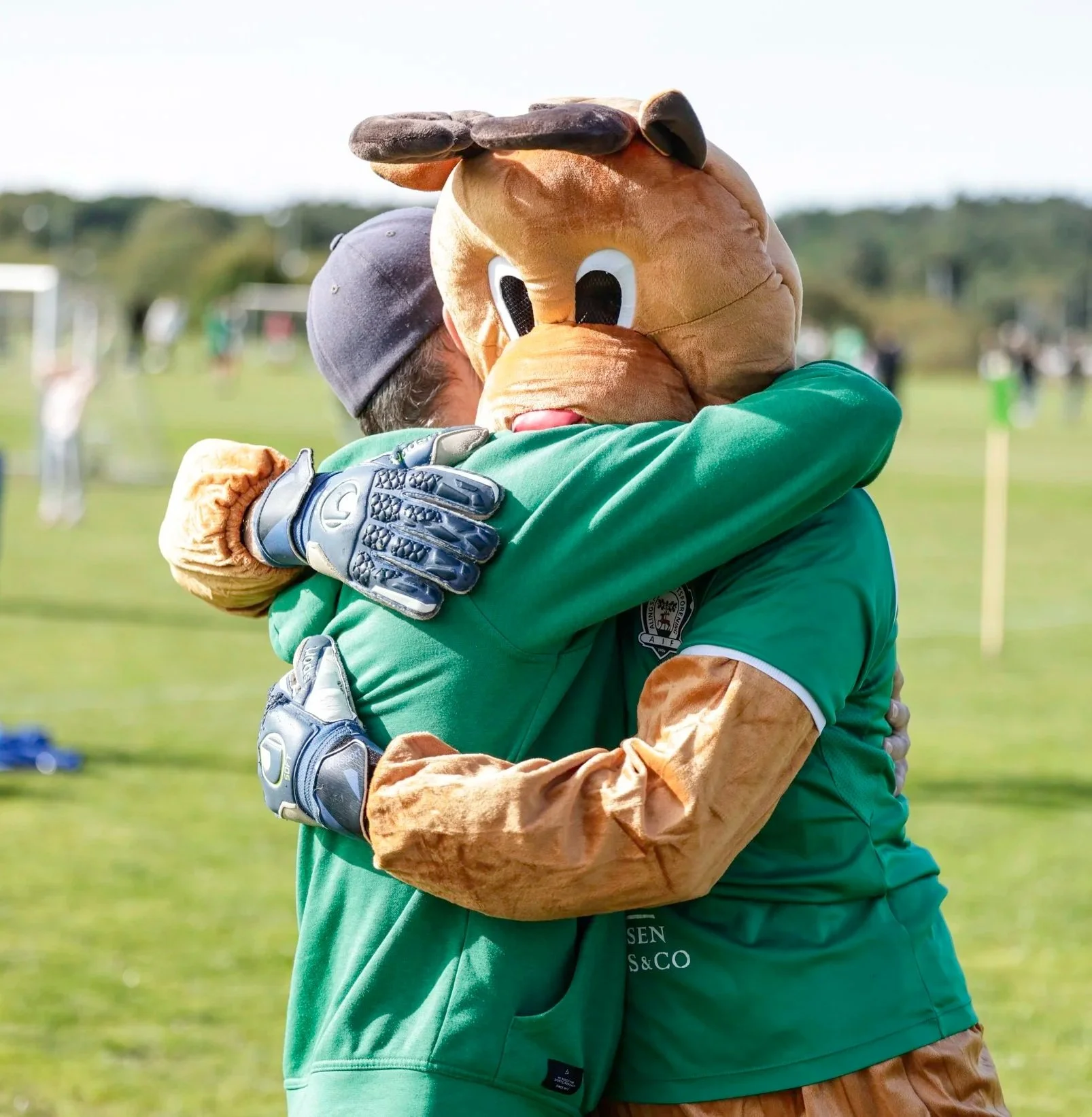 Två personer i fotbollsutrustning och en maskot som är en srceni som kramas på en fotbollsplan.