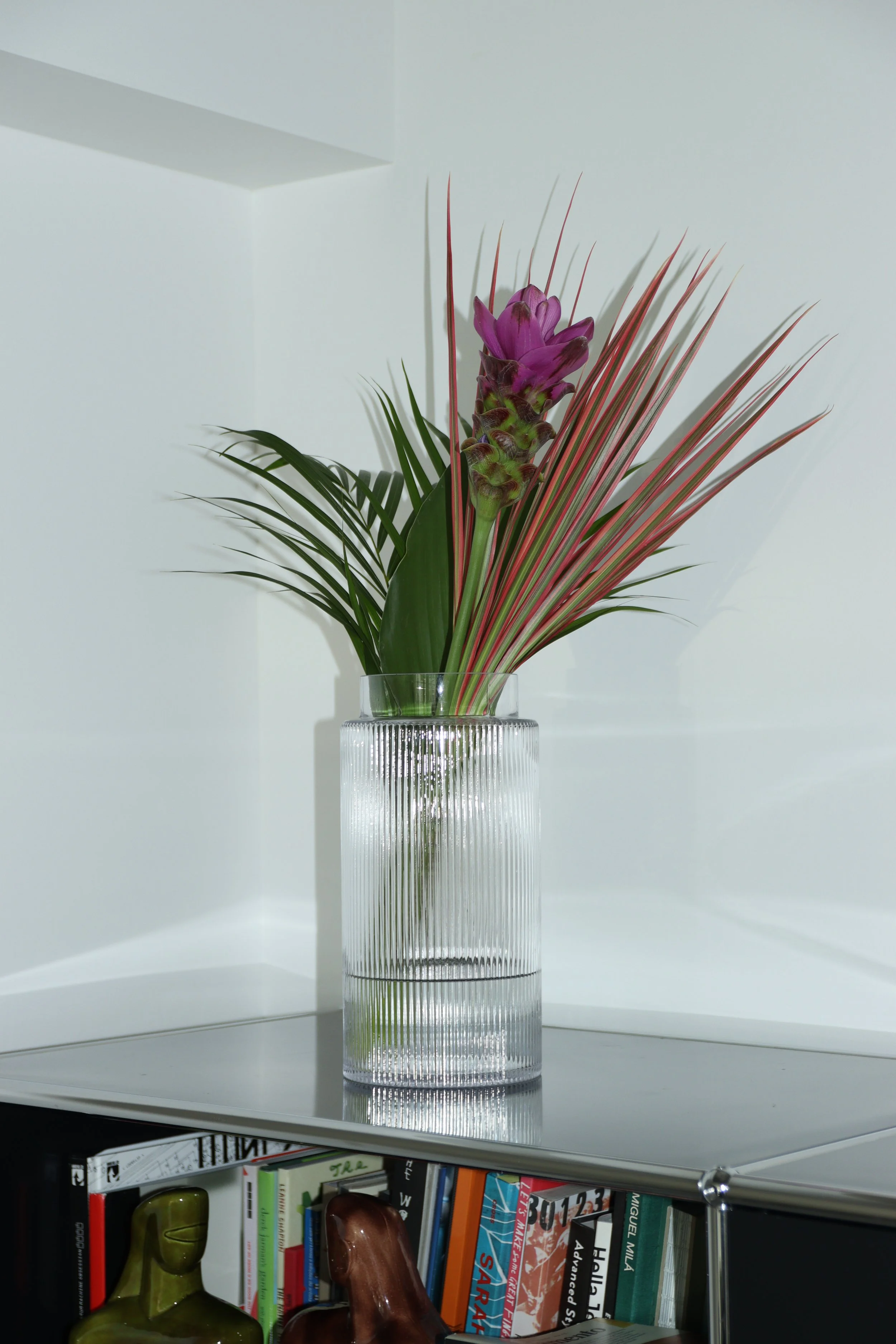 紫の花と放射状に広がる葉をガラス花器に生けた室内のフラワーアレンジメント。Indoor floral arrangement with a purple blossom and radiating foliage in a glass vase.