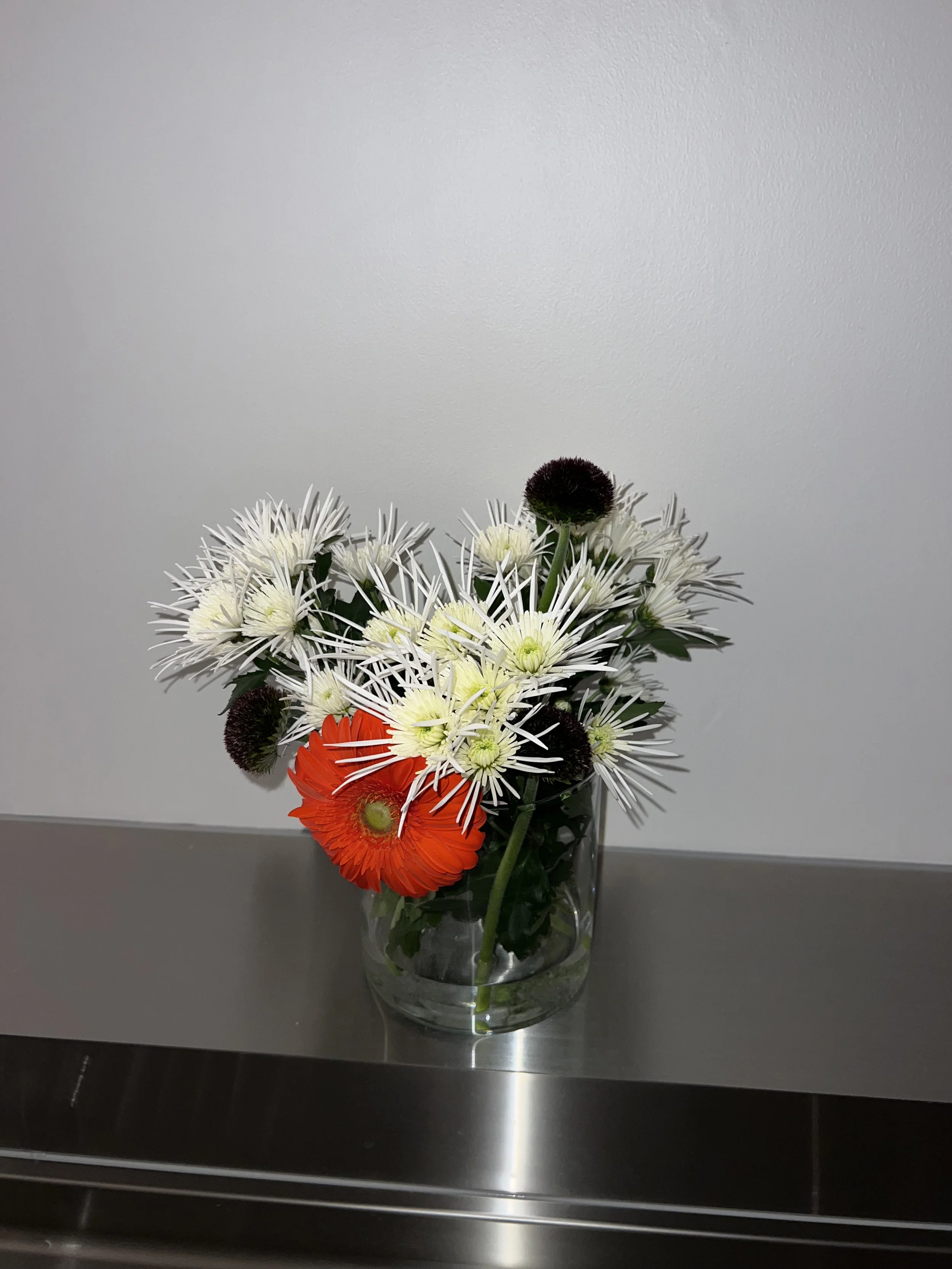 白いスパイダー咲きの花とオレンジのガーベラを組み合わせた室内のフラワーアレンジメント。Indoor floral arrangement featuring white spider chrysanthemums and an orange gerbera.