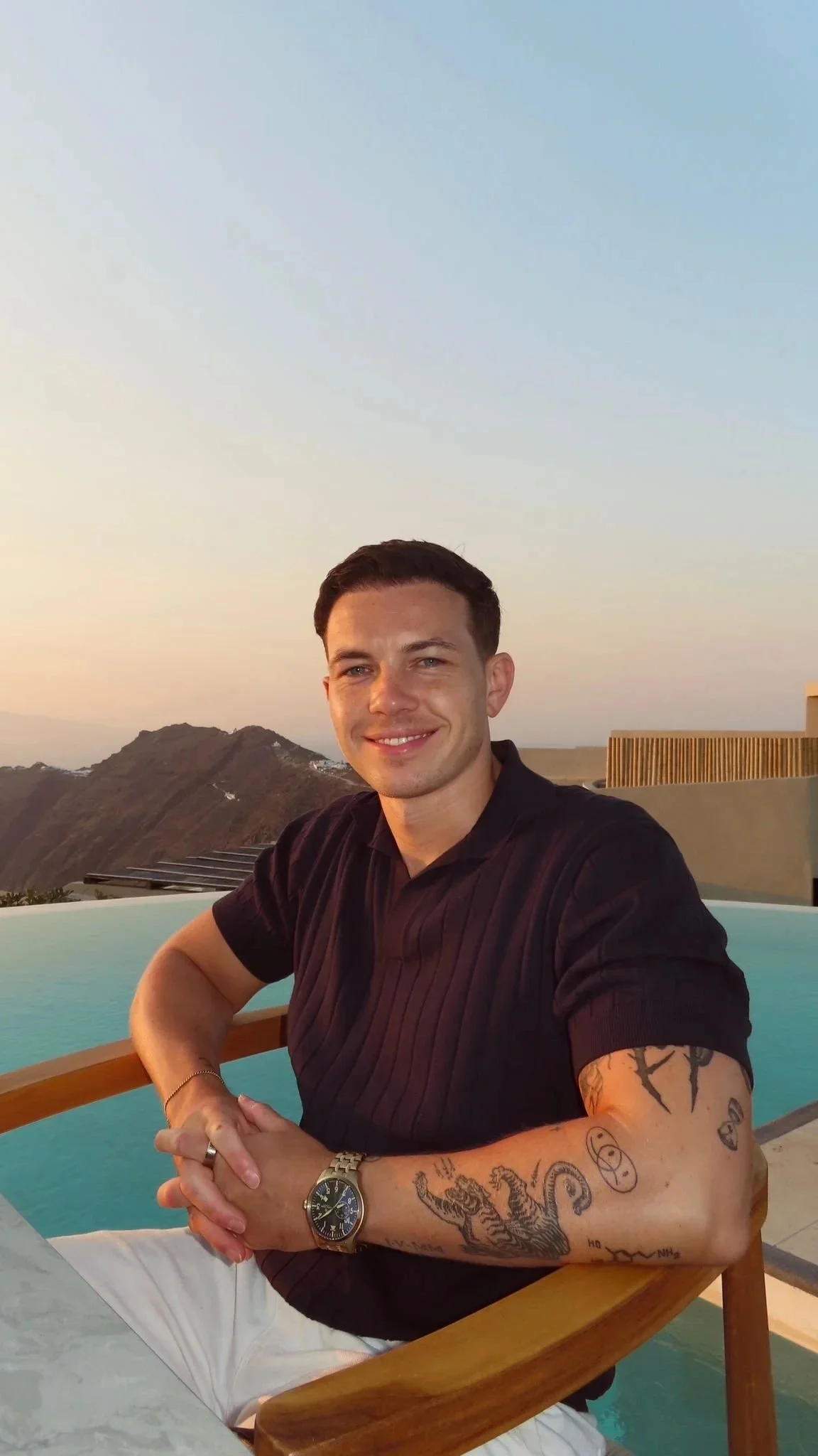 A smiling man with tattoos on his right arm, wearing a black shirt and a watch, sitting at a table near a pool with a mountain view at sunset.