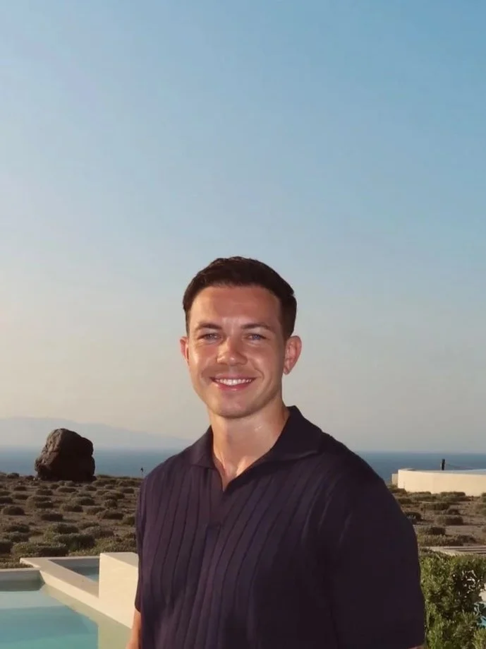 A smiling man in his late twenties with dark hair wearing a black shirt standing outdoors with a coastal view in the background.