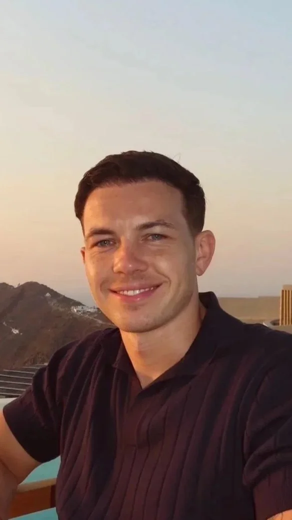 A smiling man late twenties with short dark hair and blue eyes smiling at the camera during sunset. He is wearing a black shirt and sitting outdoors with a scenic background of hills and a pastel sky.