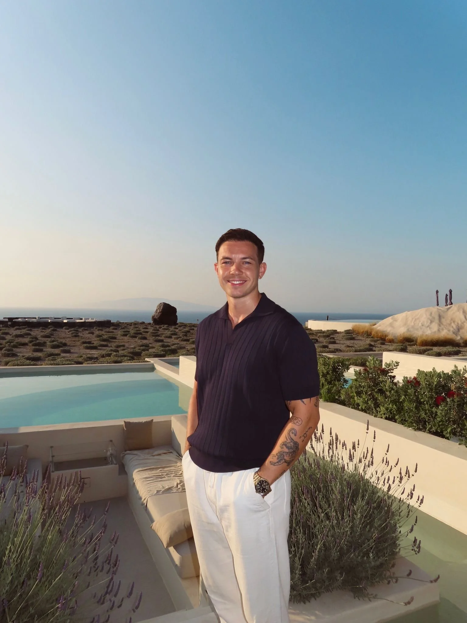 A man standing outdoors by a pool with a scenic landscape and ocean in the background, smiling and dressed in a black shirt and white pants.