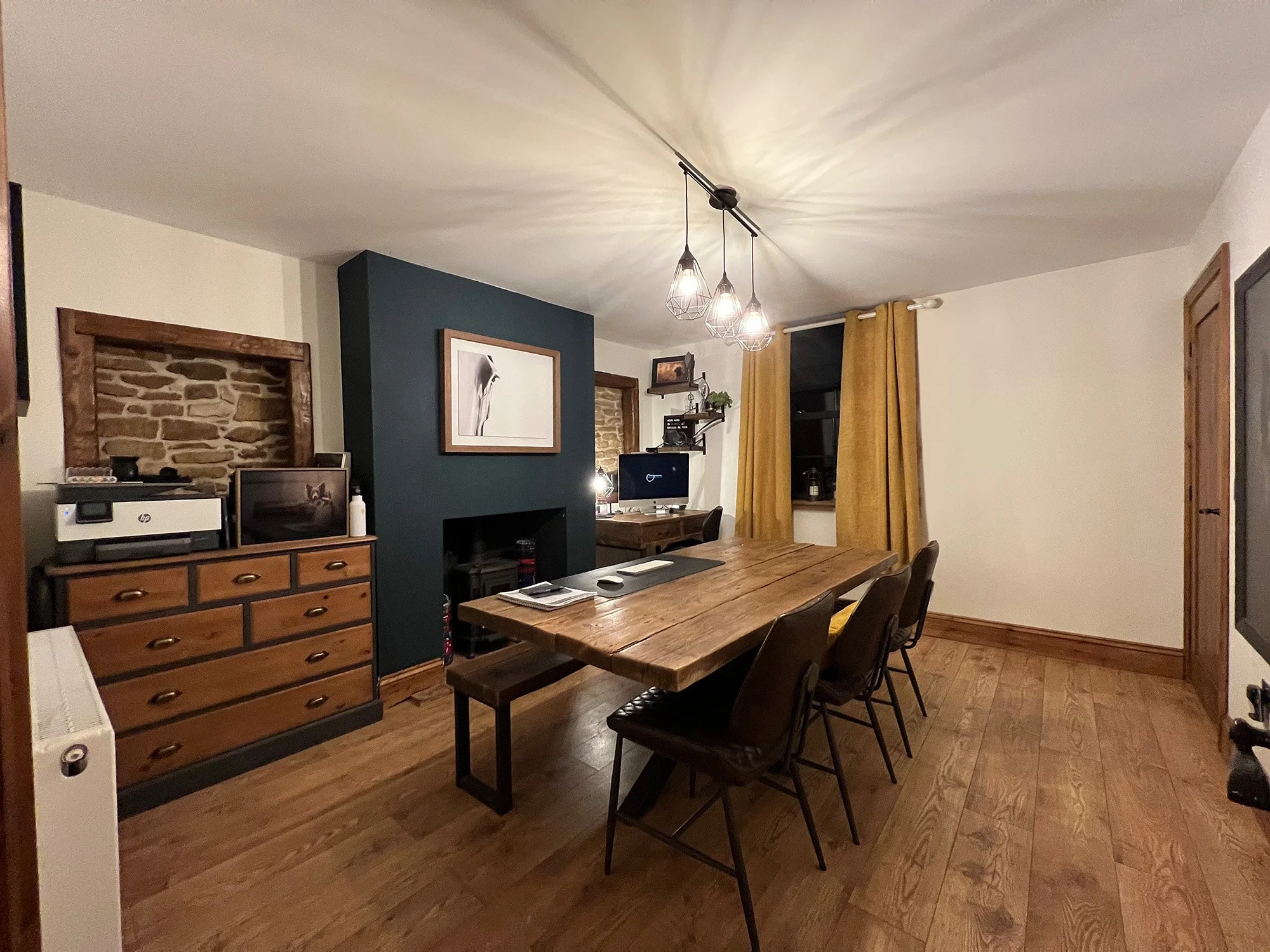 A dining room with a wooden table and six chairs, a dark green accent wall with a picture, a wooden sideboard with a printer and a monitor, a window with yellow curtains, and a ceiling light fixture.