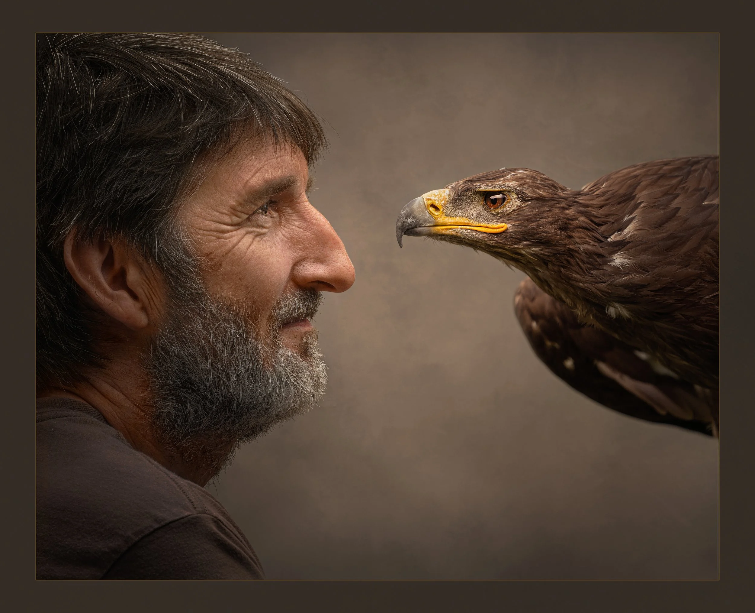 A man with gray hair and a beard facing an eagle with brown feathers and yellow beak, both staring intensely at each other.