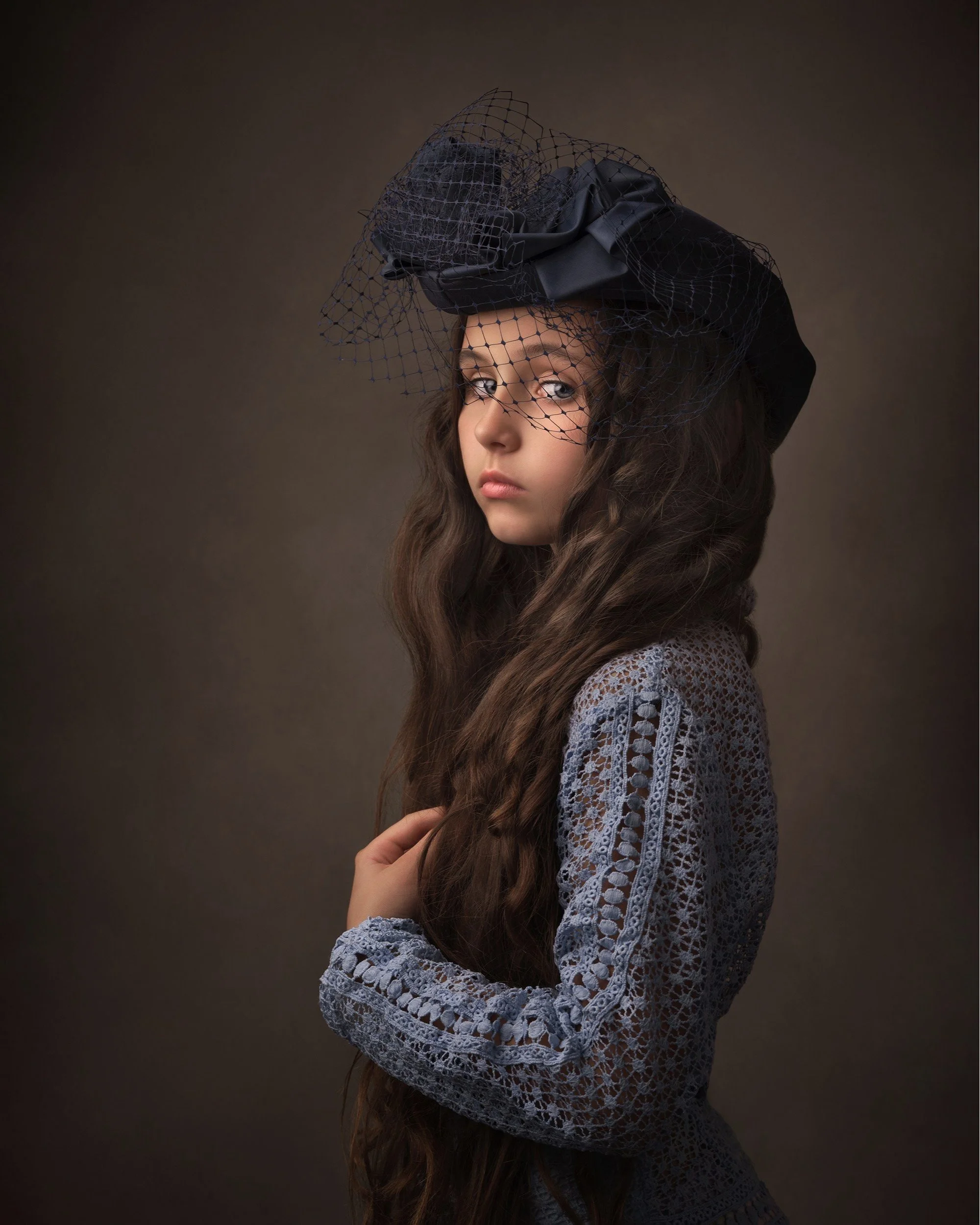 Portrait of a young woman with long brown hair wearing a black hat with a veil and a gray lace top, against a dark background.