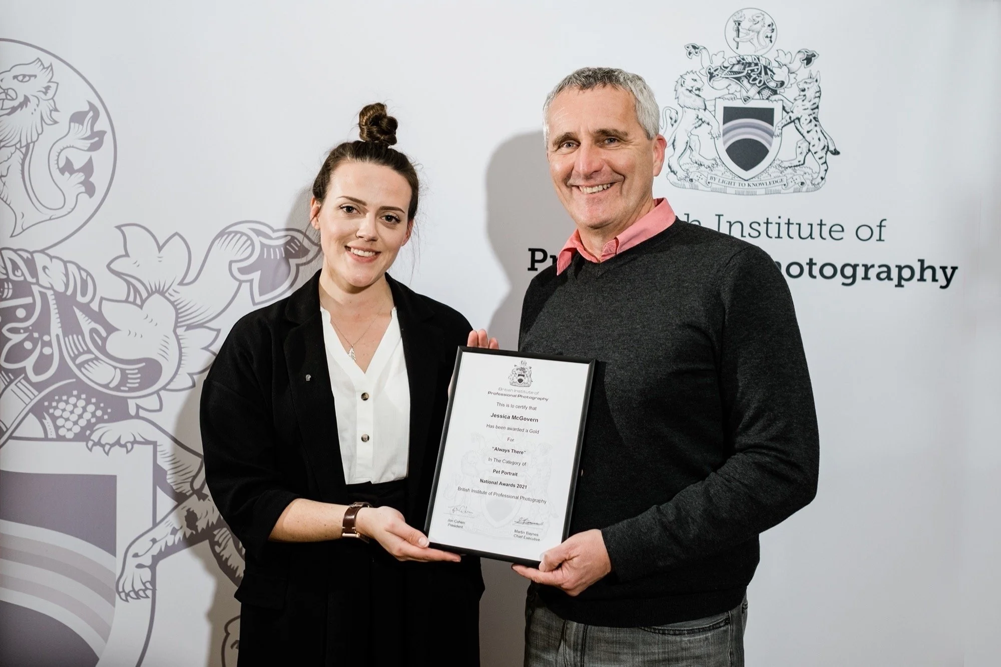 A young woman and an older man smiling and holding a framed award certificate in front of a backdrop with the logo and name of the British Institute of Professional Photography.