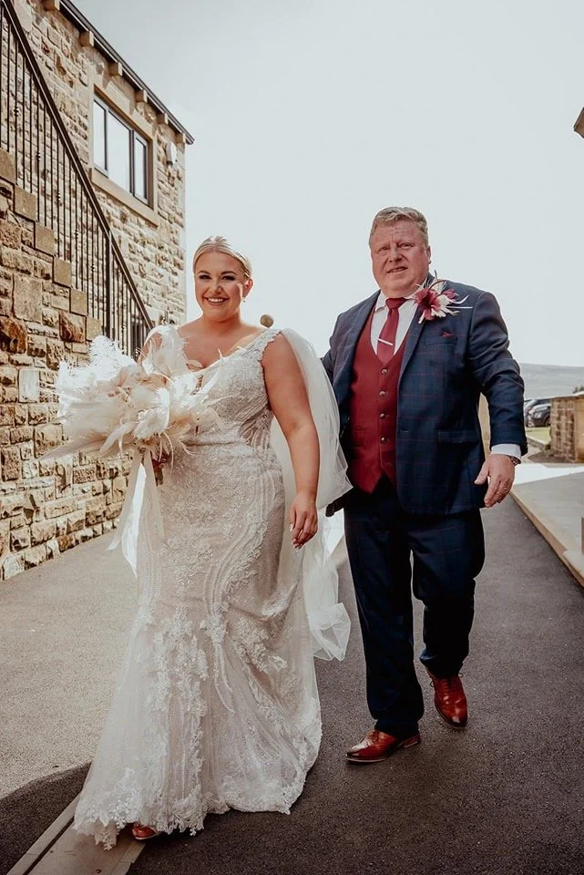 A bride in a white lace wedding gown holding a bouquet, walking beside a man in a dark suit with a red vest and tie, outside near a stone building.