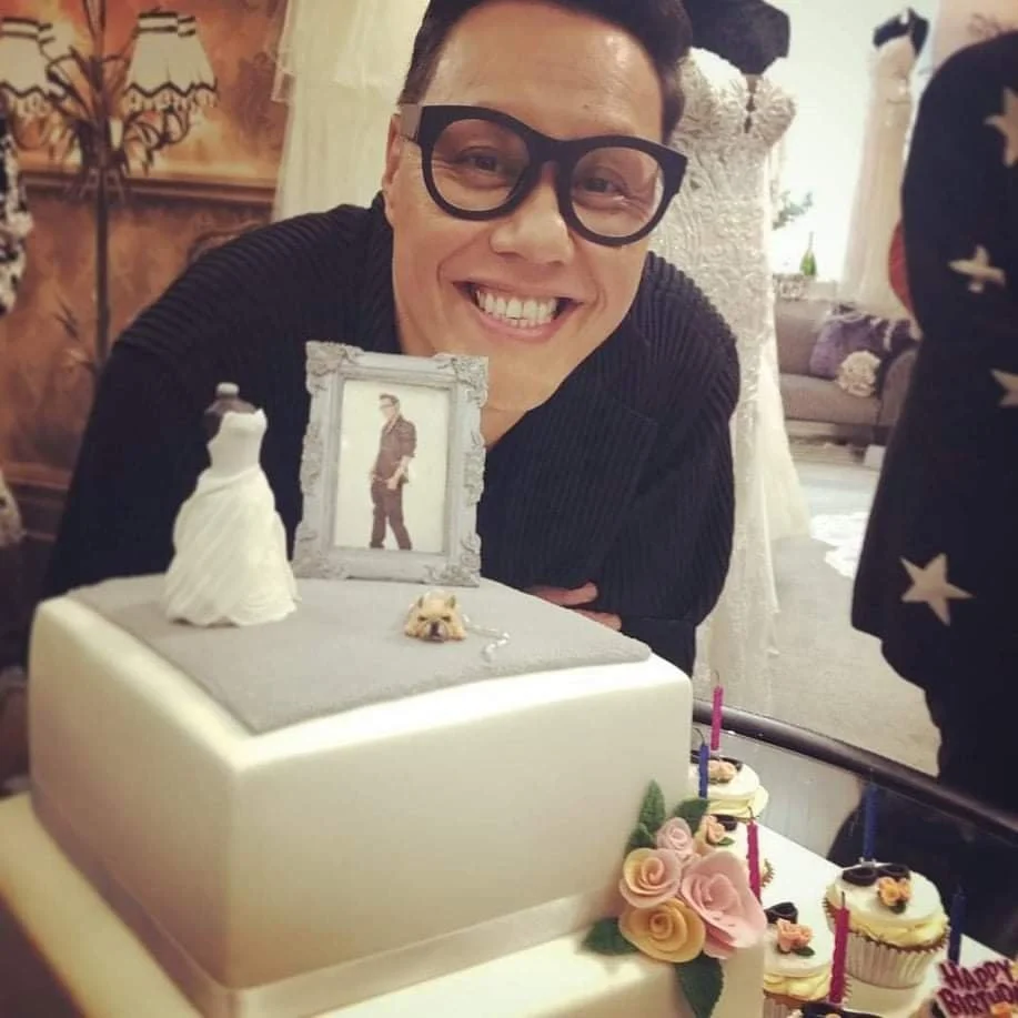 Person smiling with glasses at a cake decorated with flowers and a framed picture, in a cozy indoor setting.