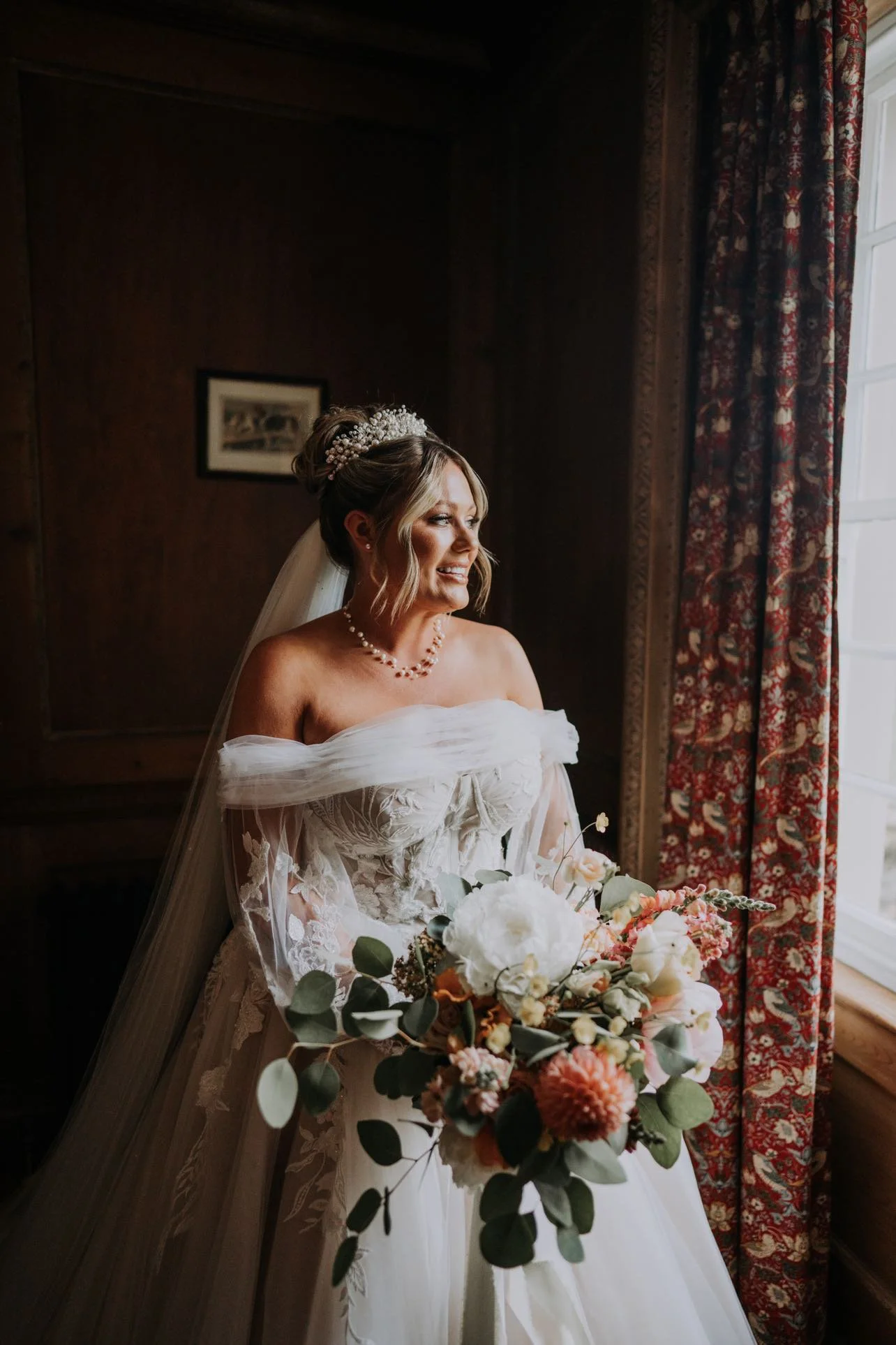 A bride in a white wedding dress with floral lace details, holding a bouquet of white and pink flowers with green leaves, standing by a window with patterned curtains, smiling as she looks outside.
