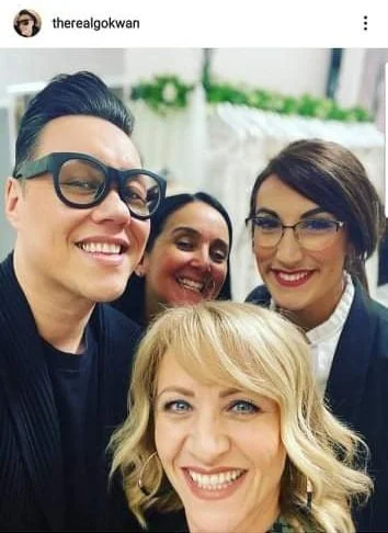 Four women taking a selfie outdoors, smiling, with greenery in the background.