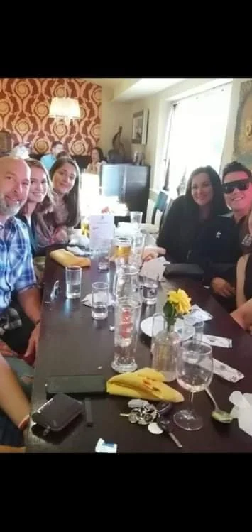 Group of six people sitting around a table in a restaurant or cafe, with drinks and tableware, smiling for the photo.