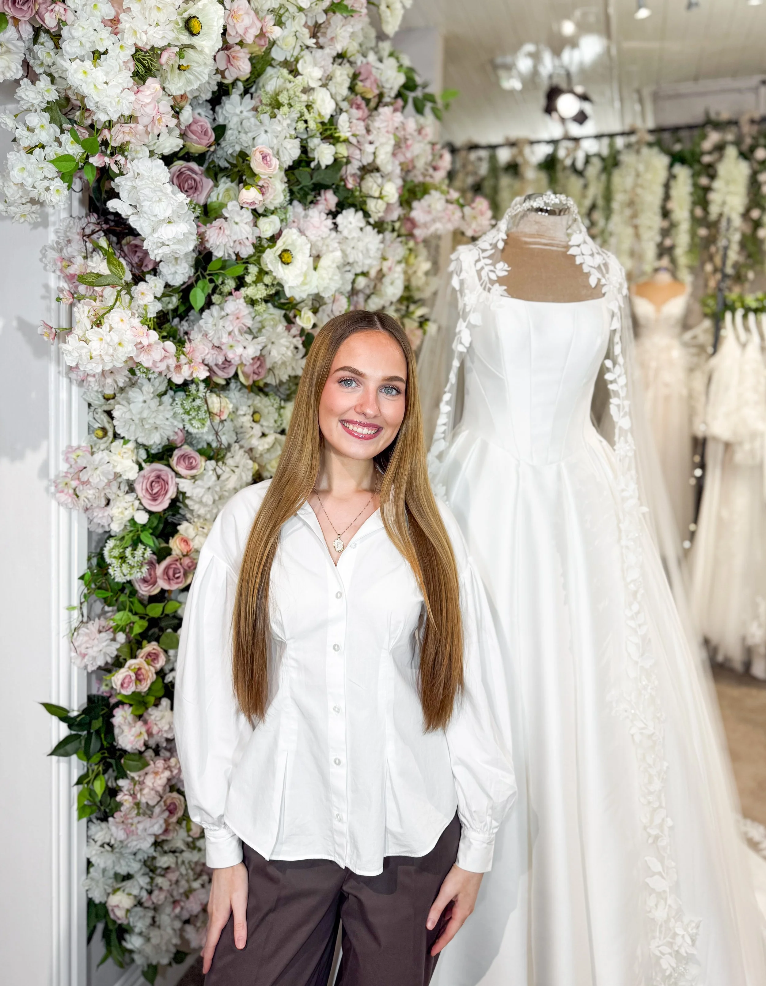 A woman with long brown hair smiling in front of wedding dresses and a floral arrangement.