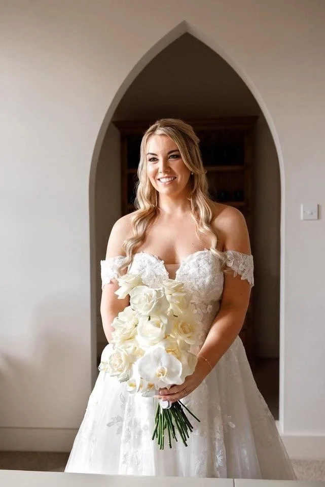 A bride in a white wedding dress holding a bouquet of white roses, standing inside a room with an arched doorway.